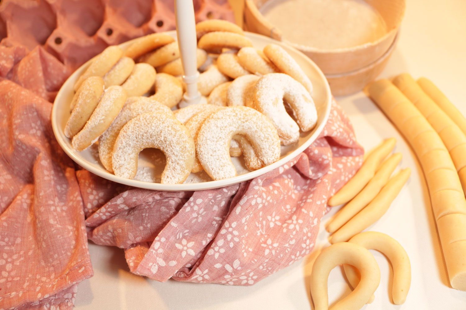 Ein Teller mit Vanillekipferln auf einem rosa Tuch, umgeben von Teigrollen.