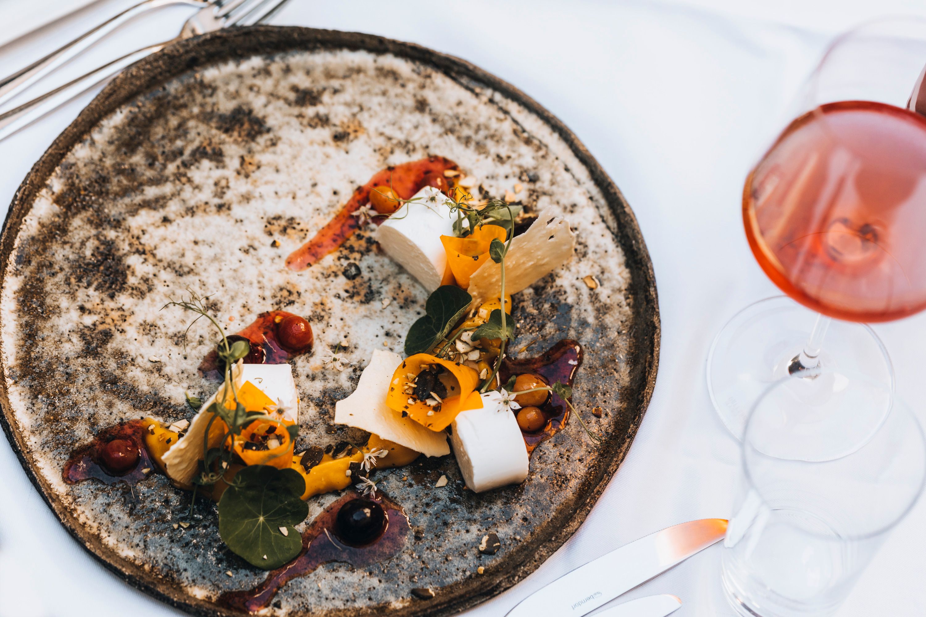 Ein kunstvoll angerichteter Teller mit mariniertem Schafkäse, Kürbis und Traubensalat, daneben ein Glas Roséwein.