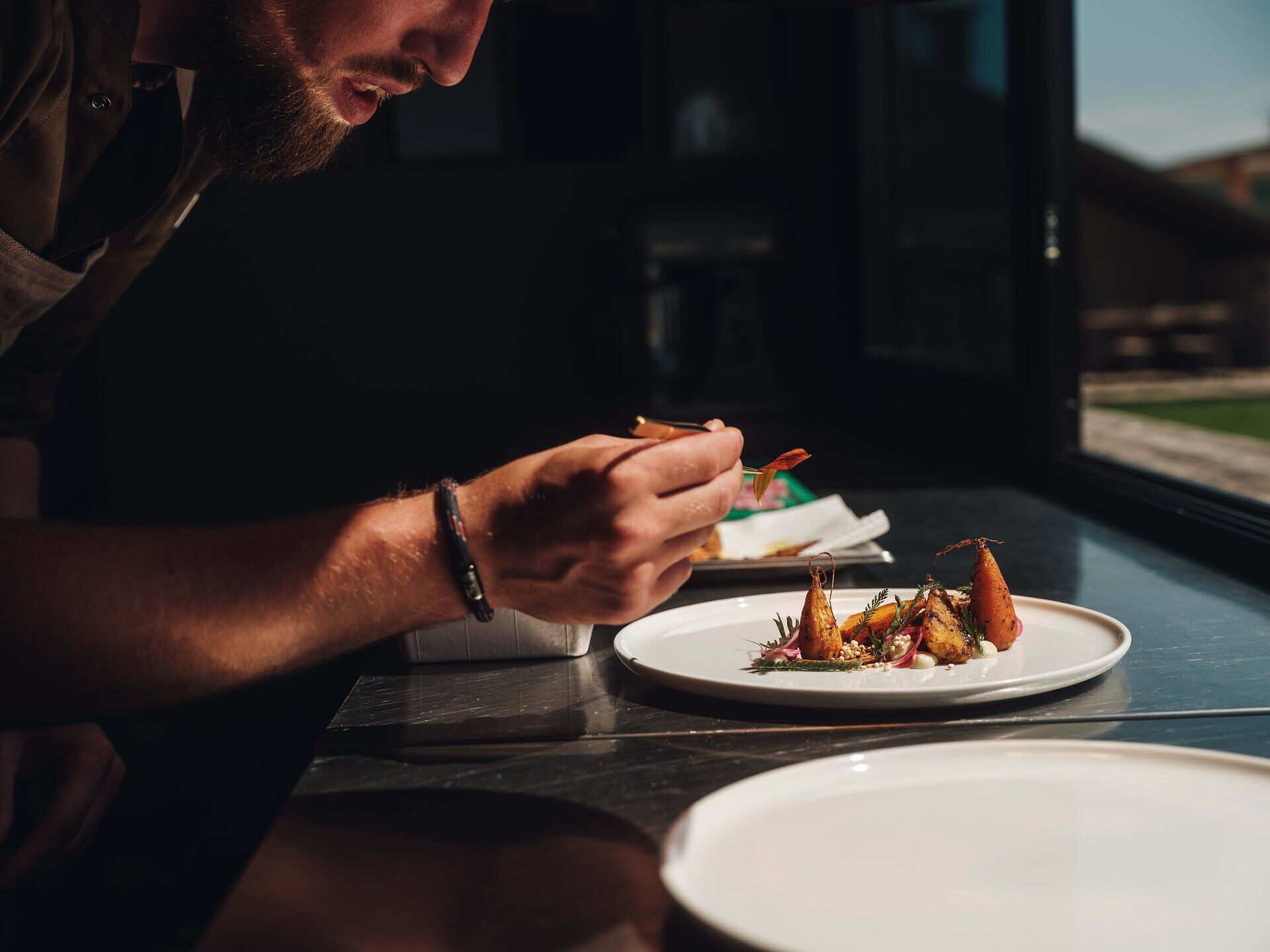 Ein leidenschaftlicher Koch konzentriert sich auf die kunstvolle Anrichtung eines exquisiten Gerichts. Die warmen Lichtstrahlen, die durch das Fenster fallen, verleihen der Szene eine einladende Atmosphäre und betonen die frischen Zutaten, die mit Liebe zum Detail präsentiert werden.