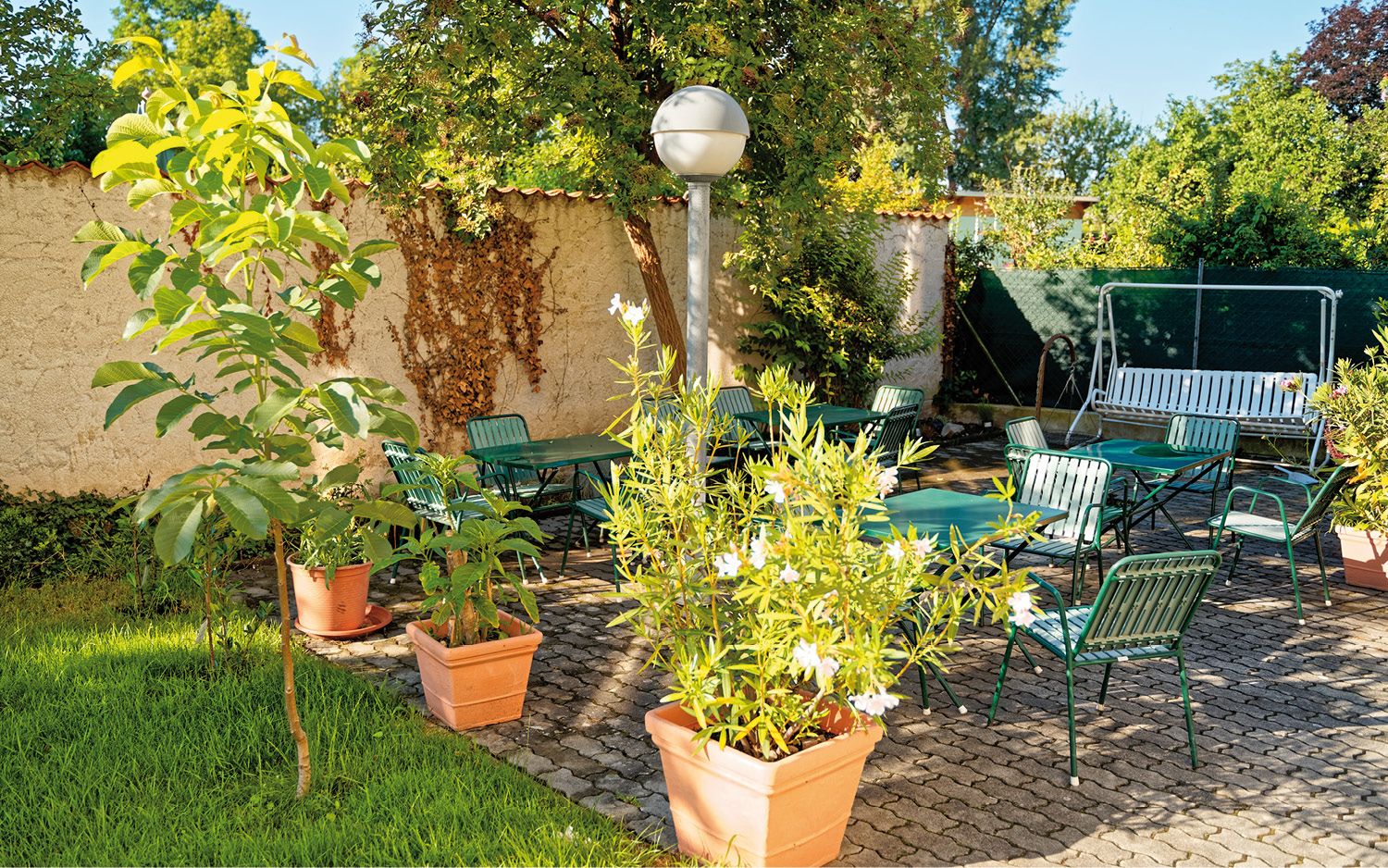Ein sonniger Garten mit grünen Stühlen, Tischen und Pflanzen in Töpfen auf einer gepflasterten Terrasse.