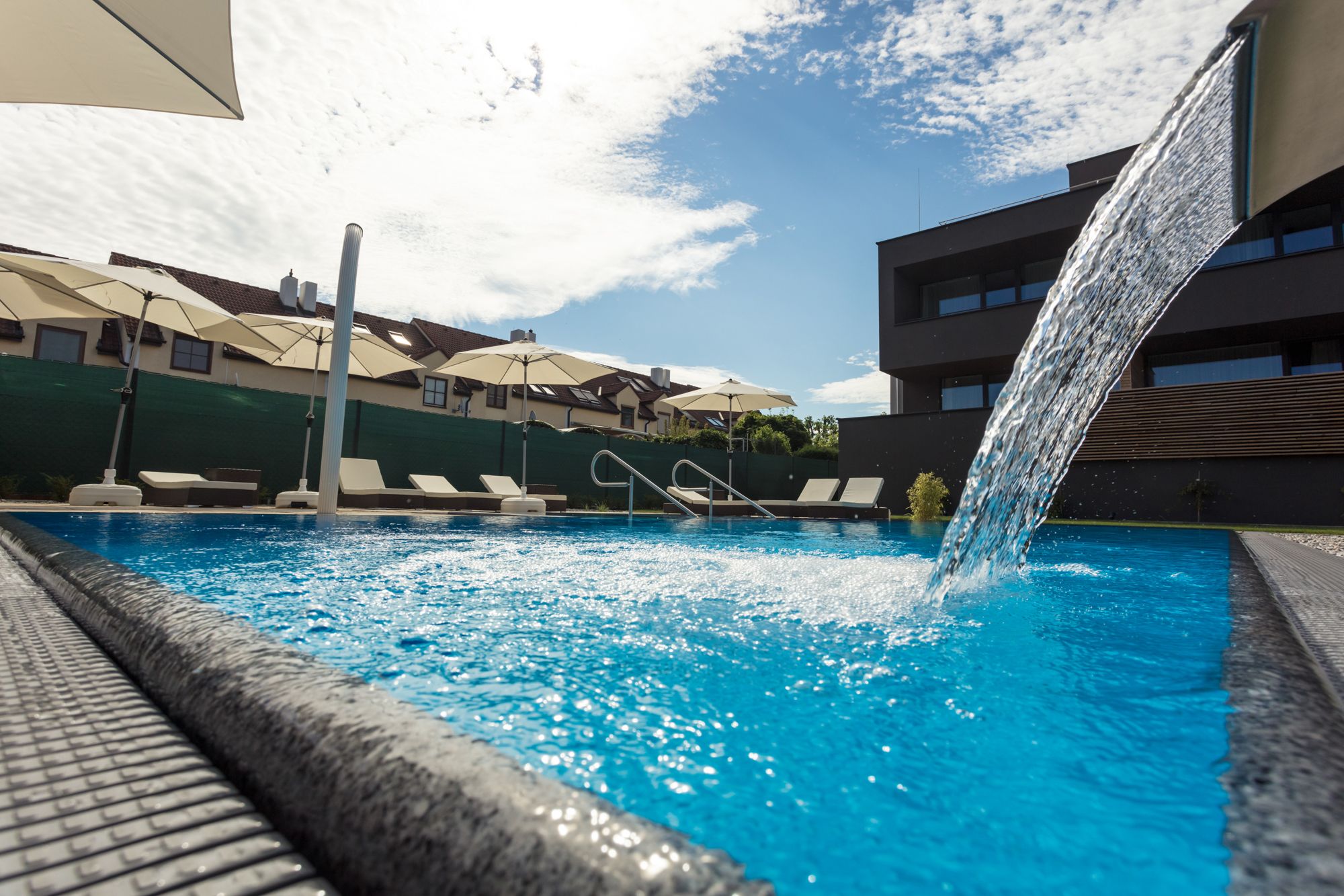 Ein moderner Pool mit Wasserfall, umgeben von Liegestühlen und Sonnenschirmen, bei sonnigem Wetter.