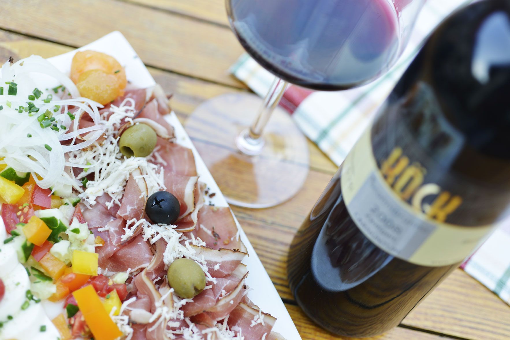 Ein Teller mit Schinken, Oliven, Gemüse und ein Glas Rotwein auf einem Holztisch.