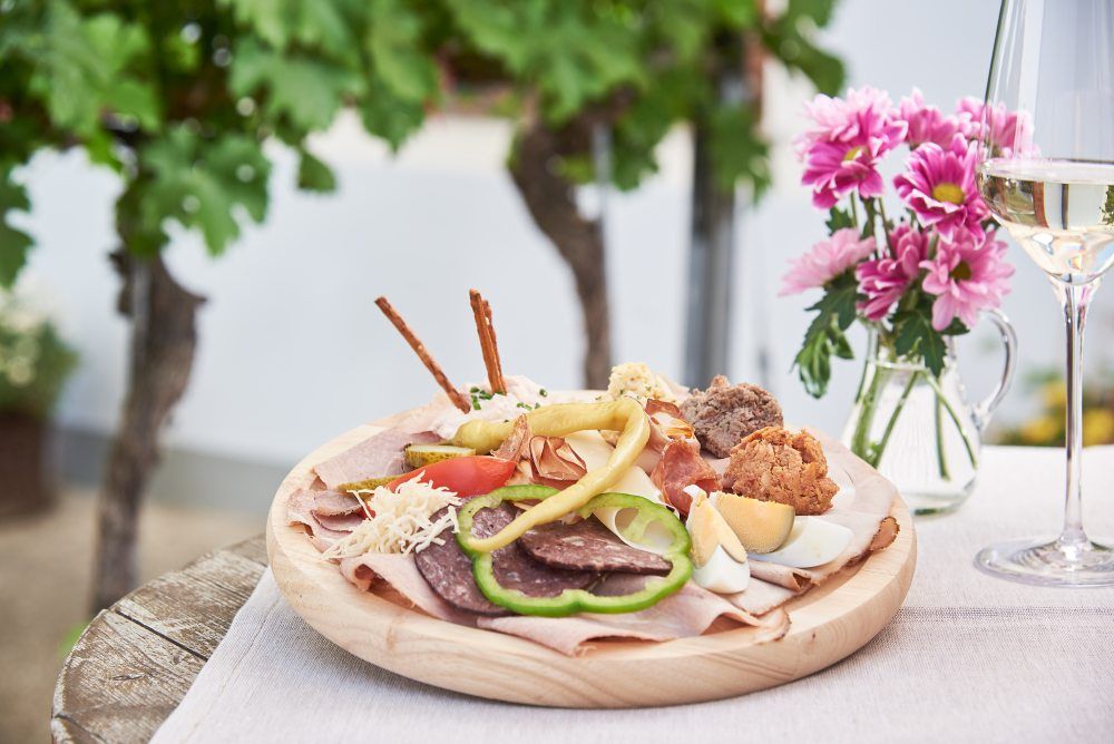 Ein Teller mit verschiedenen Heurigen-Spezialitäten, daneben ein Glas Weißwein und rosa Blumen auf einem Tisch im Freien.
