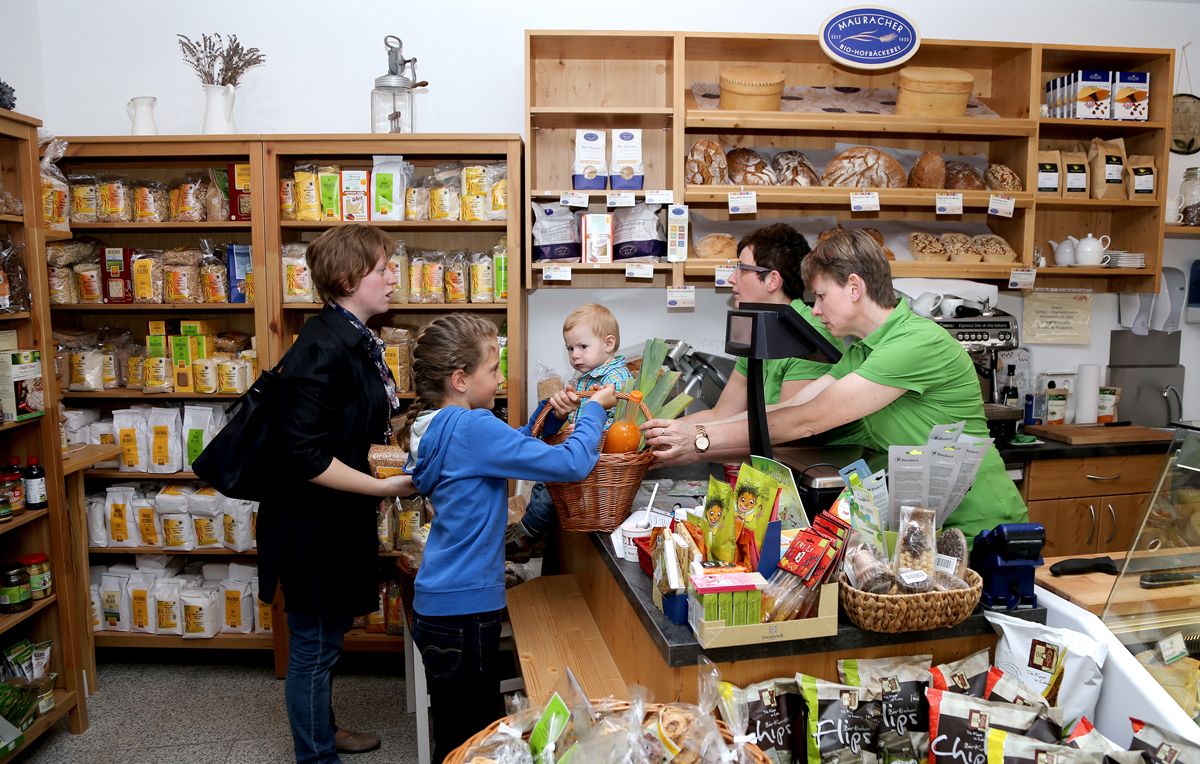 Ein kleiner Laden mit Kunden und Verkäuferinnen, die an der Theke stehen. Regale voller Bio-Produkte im Hintergrund.