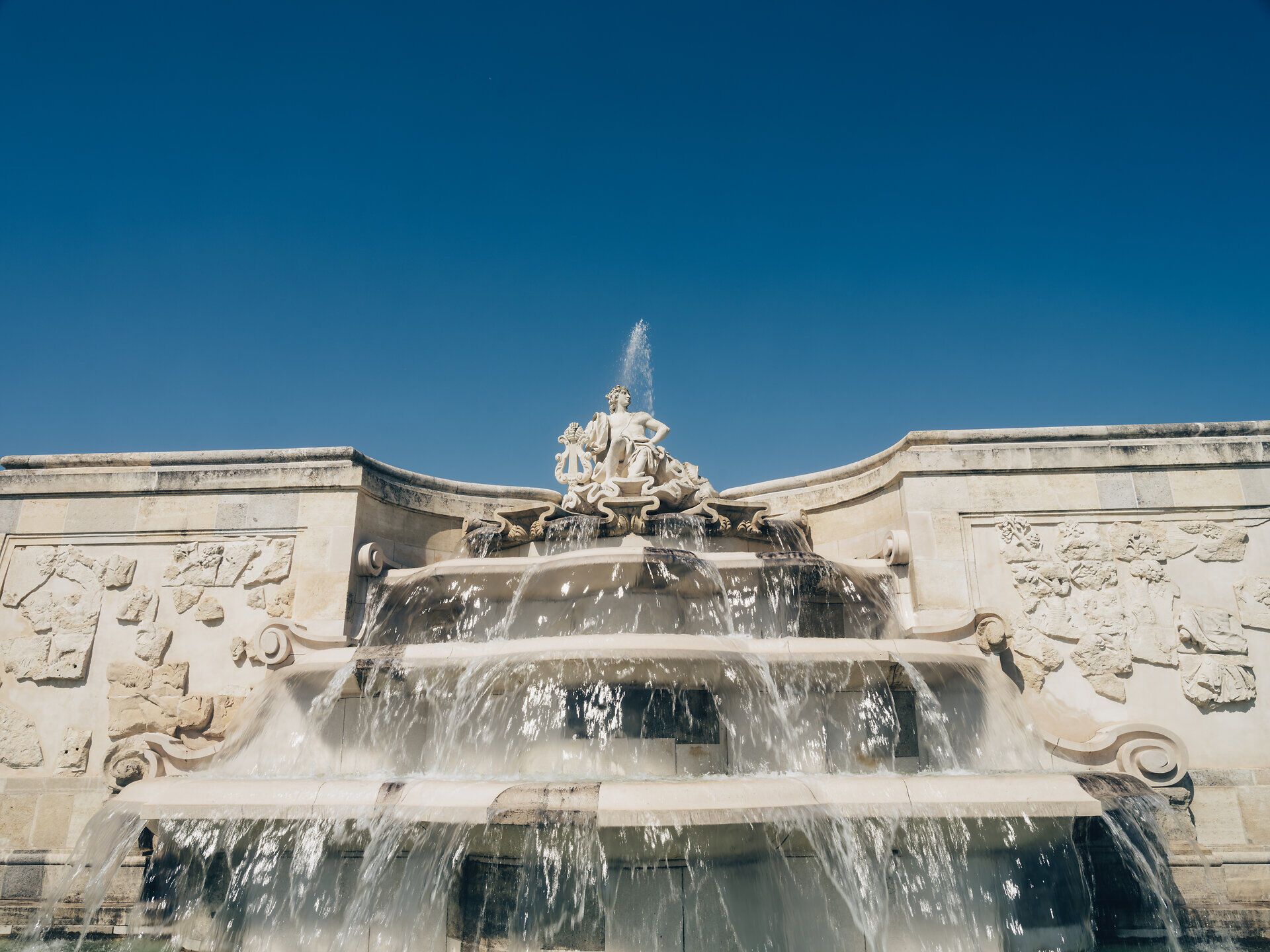 Die majestätische Fontäne sprudelt lebhaft in der warmen Sonne und verleiht dem Schloss Hof eine zauberhafte Atmosphäre. Umgeben von kunstvoll gestalteten Steinreliefs und dem klaren blauen Himmel, lädt dieser Ort zum Verweilen und Träumen ein.
