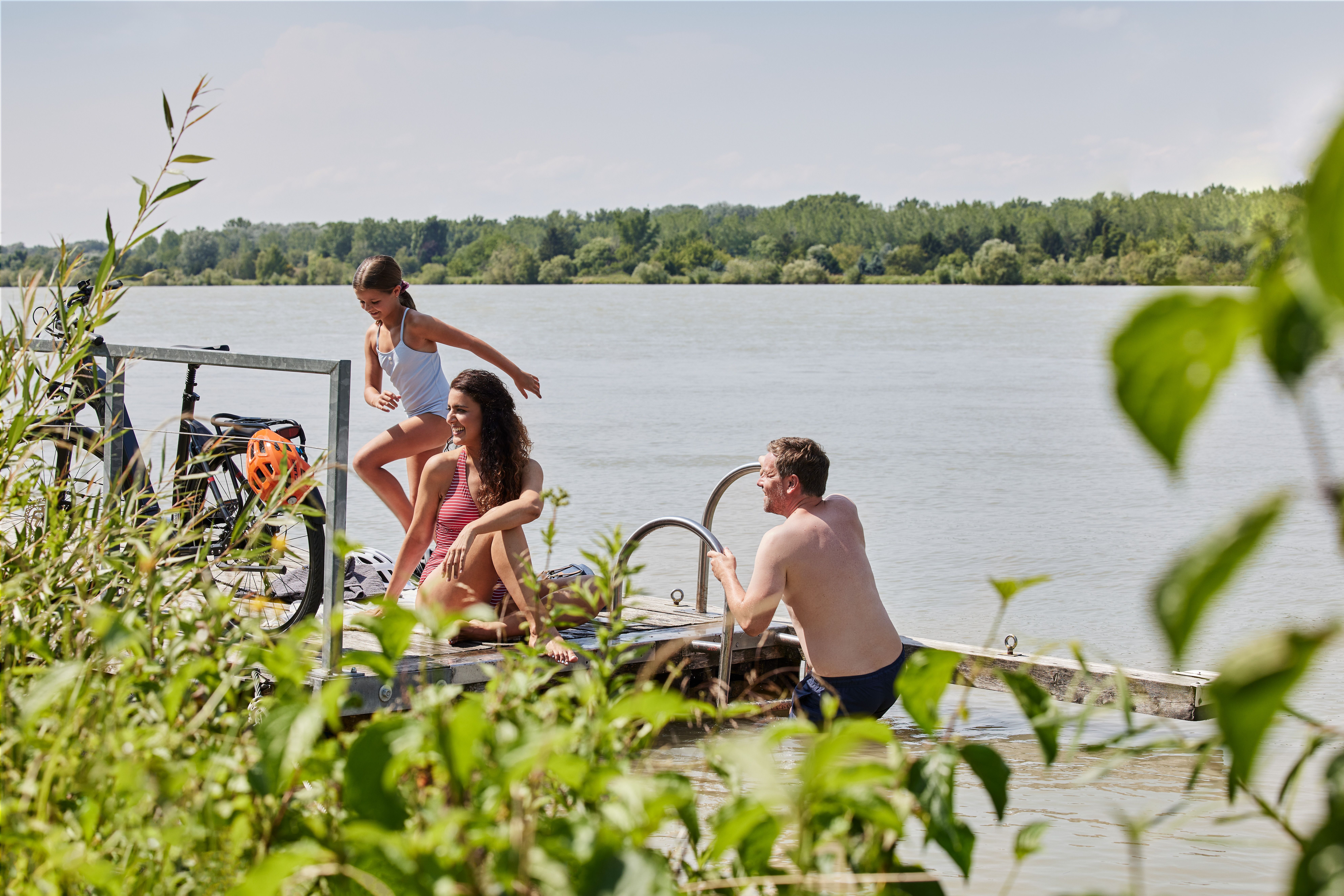 An einem sonnigen Sommertag genießen Familien die erfrischende Abkühlung im klaren Wasser der Donau. Kinder lachen und springen fröhlich vom Steg ins Wasser, während die sanfte Brise durch das grüne Uferrauschen weht. Diese idyllische Szenerie lädt dazu ein, die Schönheit der Natur und die Freude am gemeinsamen Erleben zu feiern.