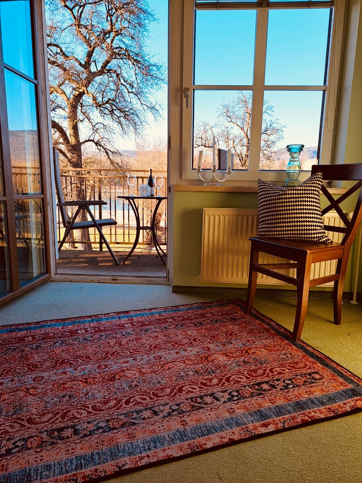 Zimmer mit Teppich, Stuhl und Blick auf einen Balkon mit Tisch und Stühlen, Bäume im Hintergrund.