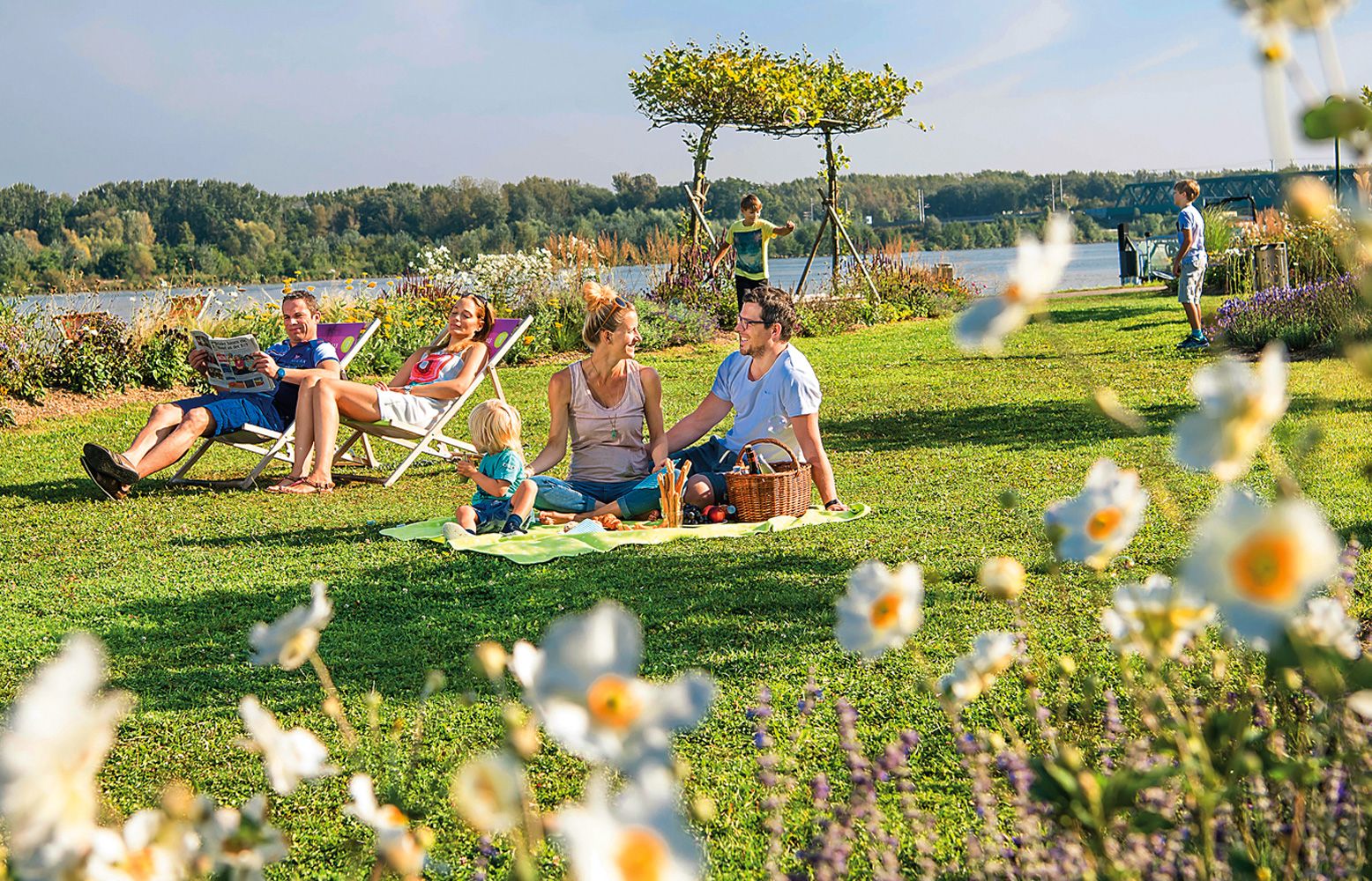 Familienpicknick im Grünen mit Blumen im Vordergrund und einem Fluss im Hintergrund.