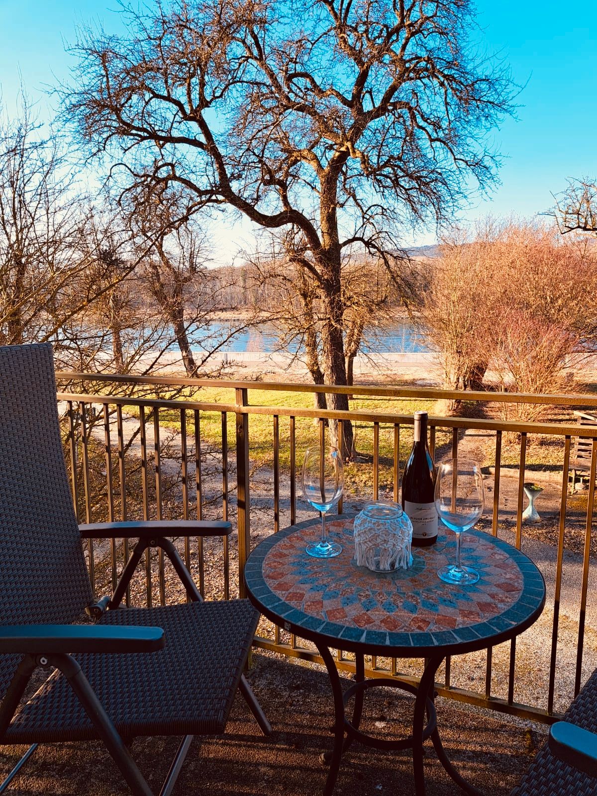 Balkon mit Tisch, zwei Weingläsern und Flasche, Blick auf Baum und Fluss.
