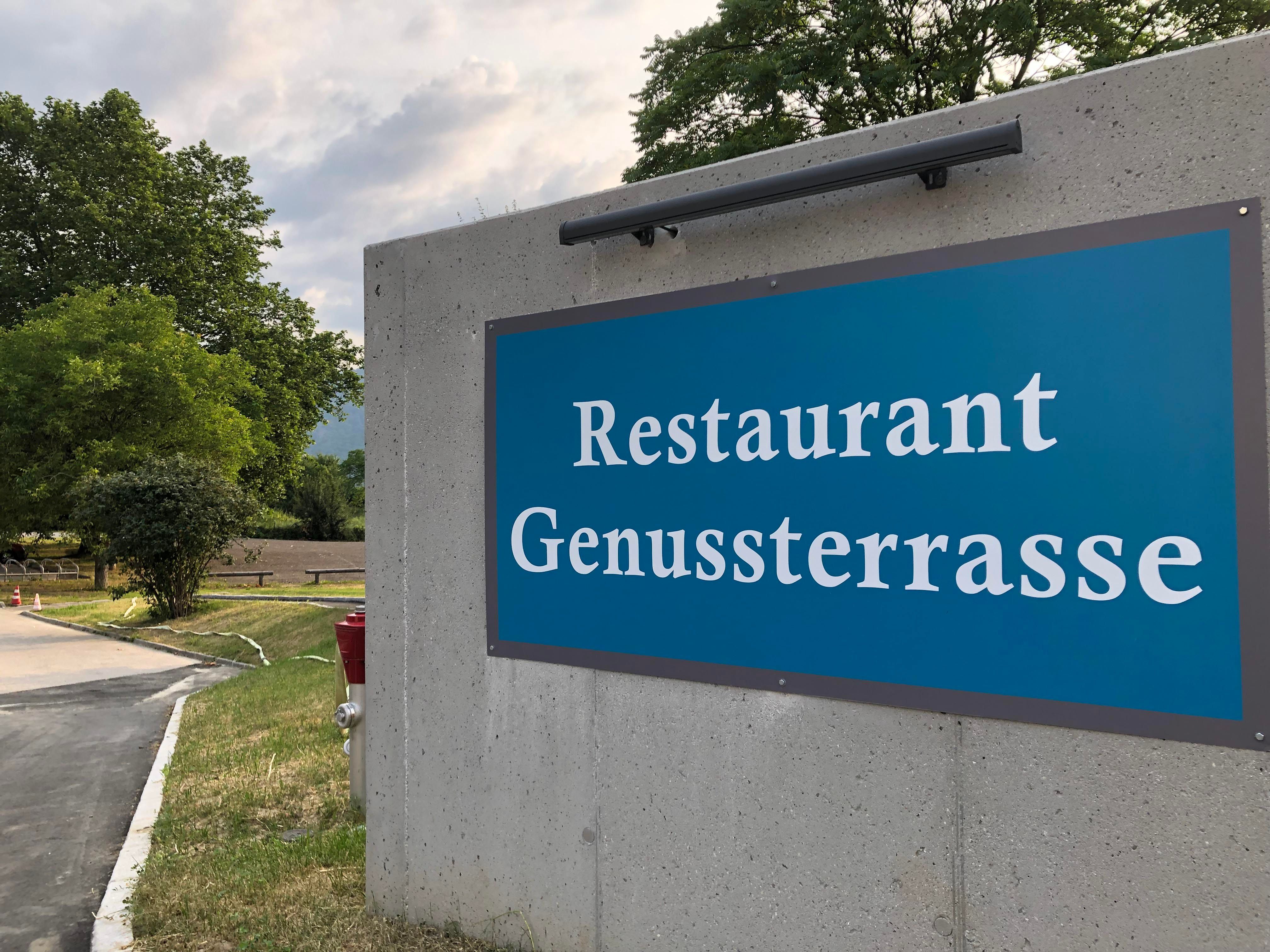 Schild des Restaurants 'Genussterrasse' an einer Betonwand im Freien.