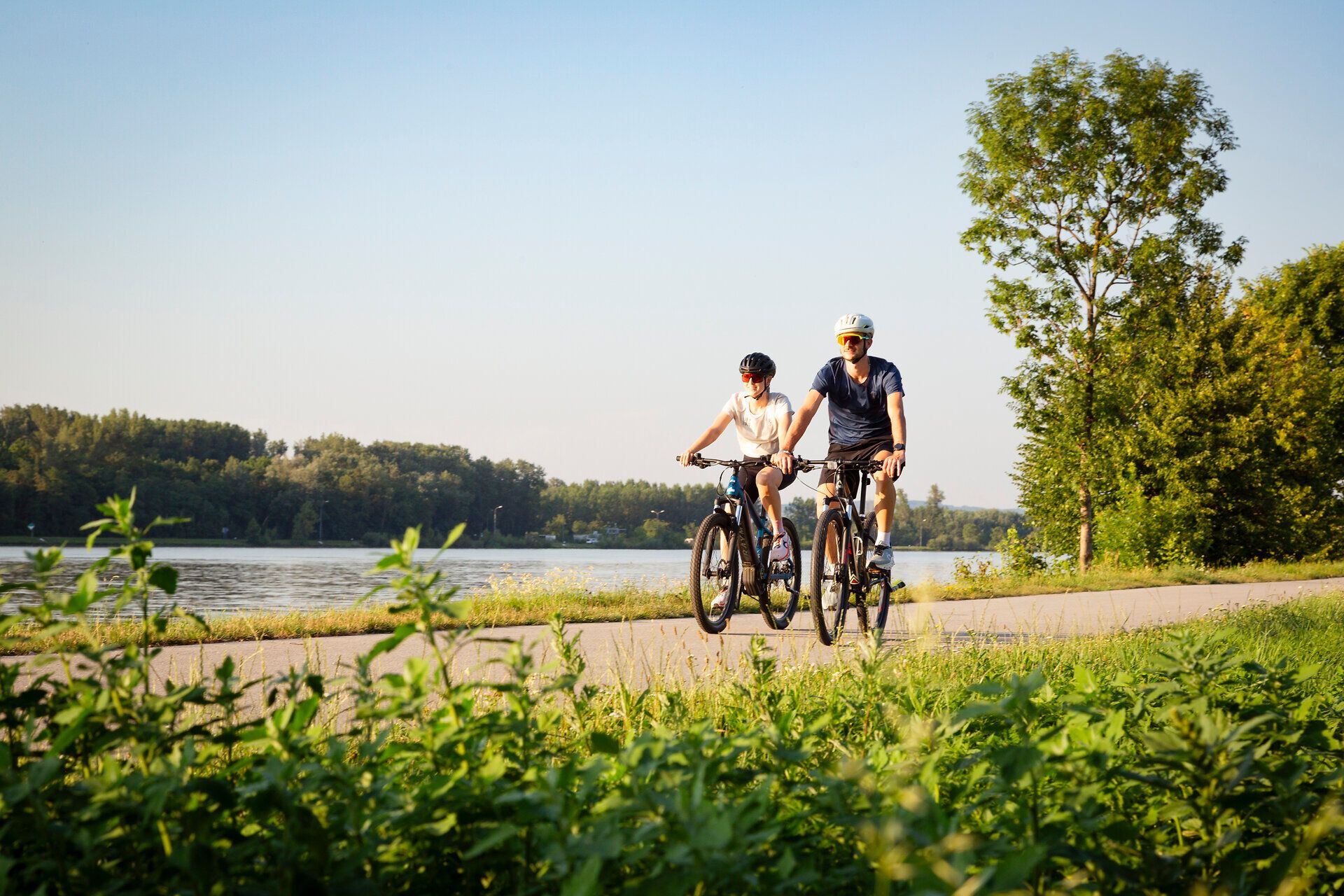 Zwei Radfahrer genießen die sanfte Brise und die malerische Aussicht entlang des Donauradwegs. Umgeben von üppigem Grün und dem glitzernden Wasser der Donau, vermittelt die Szenerie ein Gefühl von Freiheit und Abenteuer. Diese idyllische Umgebung lädt dazu ein, die Natur aktiv zu erleben und die Schönheit der Landschaft zu entdecken.