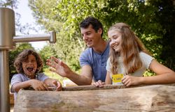 Ein fröhliches Zusammensein von Kindern und Erwachsenen am Wasser, umgeben von üppigem Grün. Das plätschernde Wasser und die strahlenden Gesichter schaffen eine einladende Atmosphäre, die zum Spielen und Entdecken einlädt.