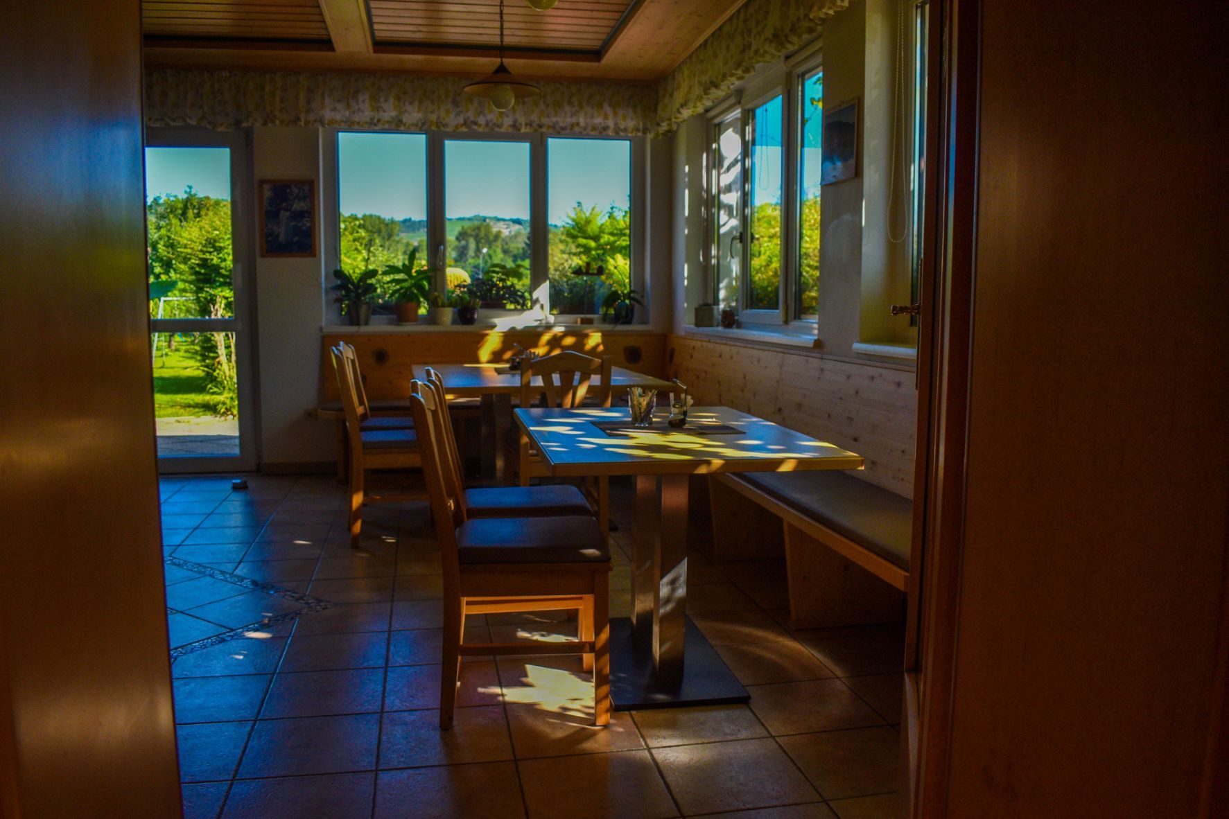 Ein gemütlicher Frühstücksraum mit Holzmöbeln und großen Fenstern, durch die Sonnenlicht fällt.