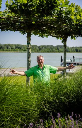 Ein Gärtner in Tulln steht hinter einem grünen Strauch, und vor einem Baum, die Donau, blauer Himmel und Sonnenschein sind zu sehen.
