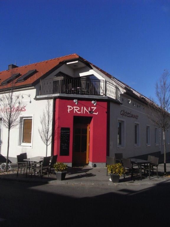 Außenansicht eines Gebäudes mit der Aufschrift 'Prinz', Gastgarten mit Tischen und Stühlen, blauer Himmel.