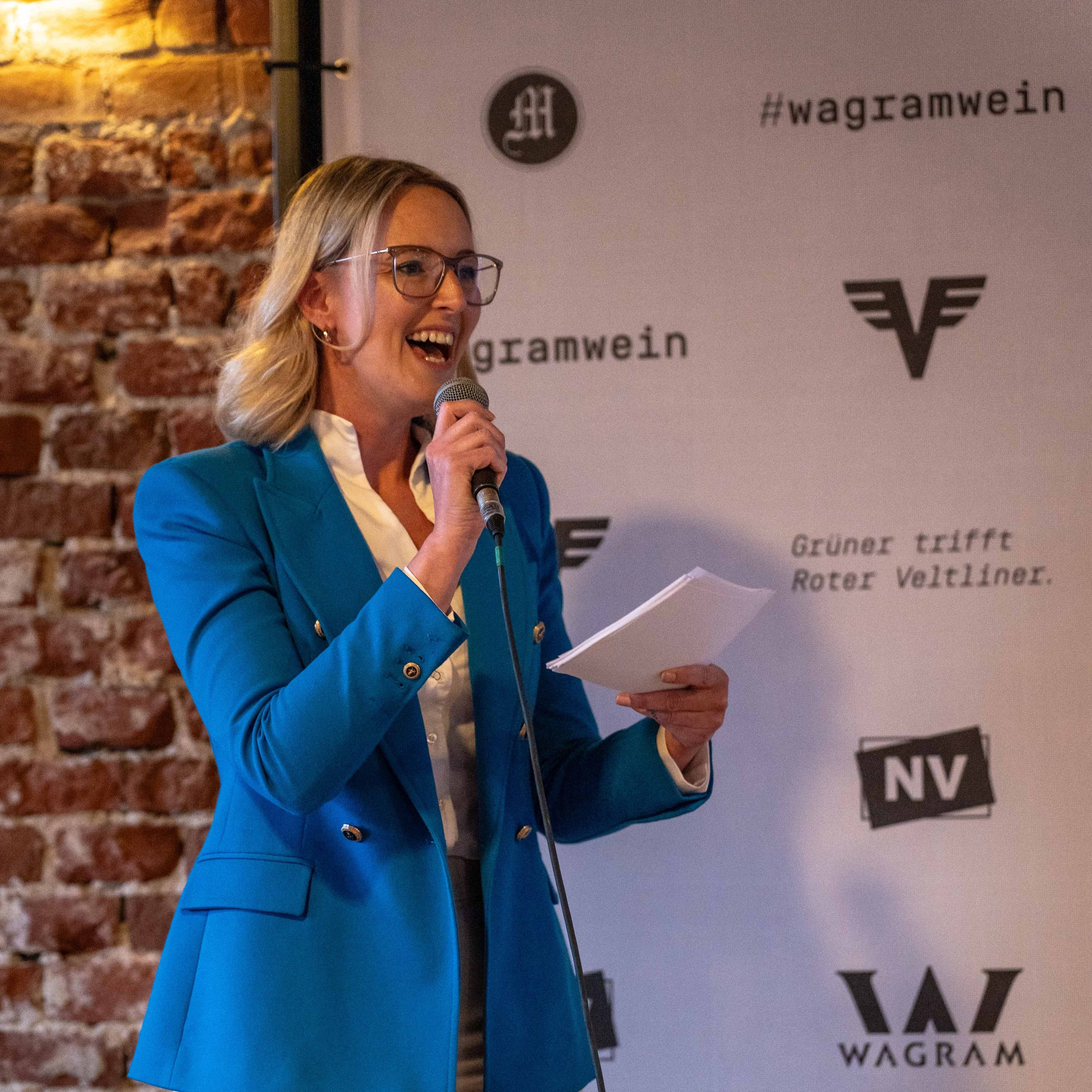 Moderatorin mit Mikrofon spricht bei einer Weinveranstaltung vor einer Wagram-Wein-Eventwand mit Logos und Backsteinwand im Hintergrund.
