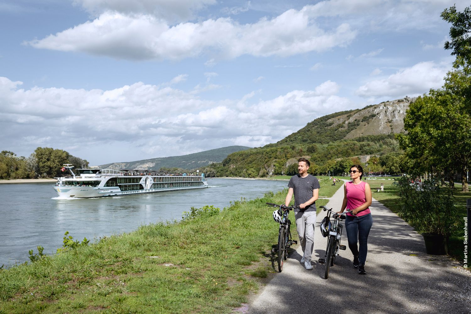 Zwei Personen schieben ein Fahrrad am Donauradweg bei Hainburg an der Donau. Im Hintergrund sieht man die Donau. 