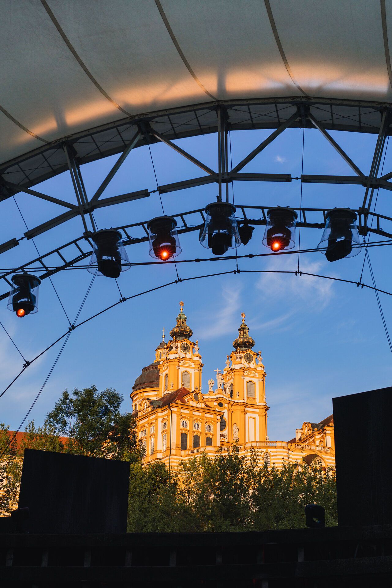 Unter dem weitläufigen Zelt des Theaterfests strahlen die warmen Lichter auf die prächtige Barockarchitektur, die im Abendlicht erblüht. Die Atmosphäre ist erfüllt von Vorfreude und der Magie des Festivals, während die Donau sanft im Hintergrund fließt und die Kulisse verzaubert.