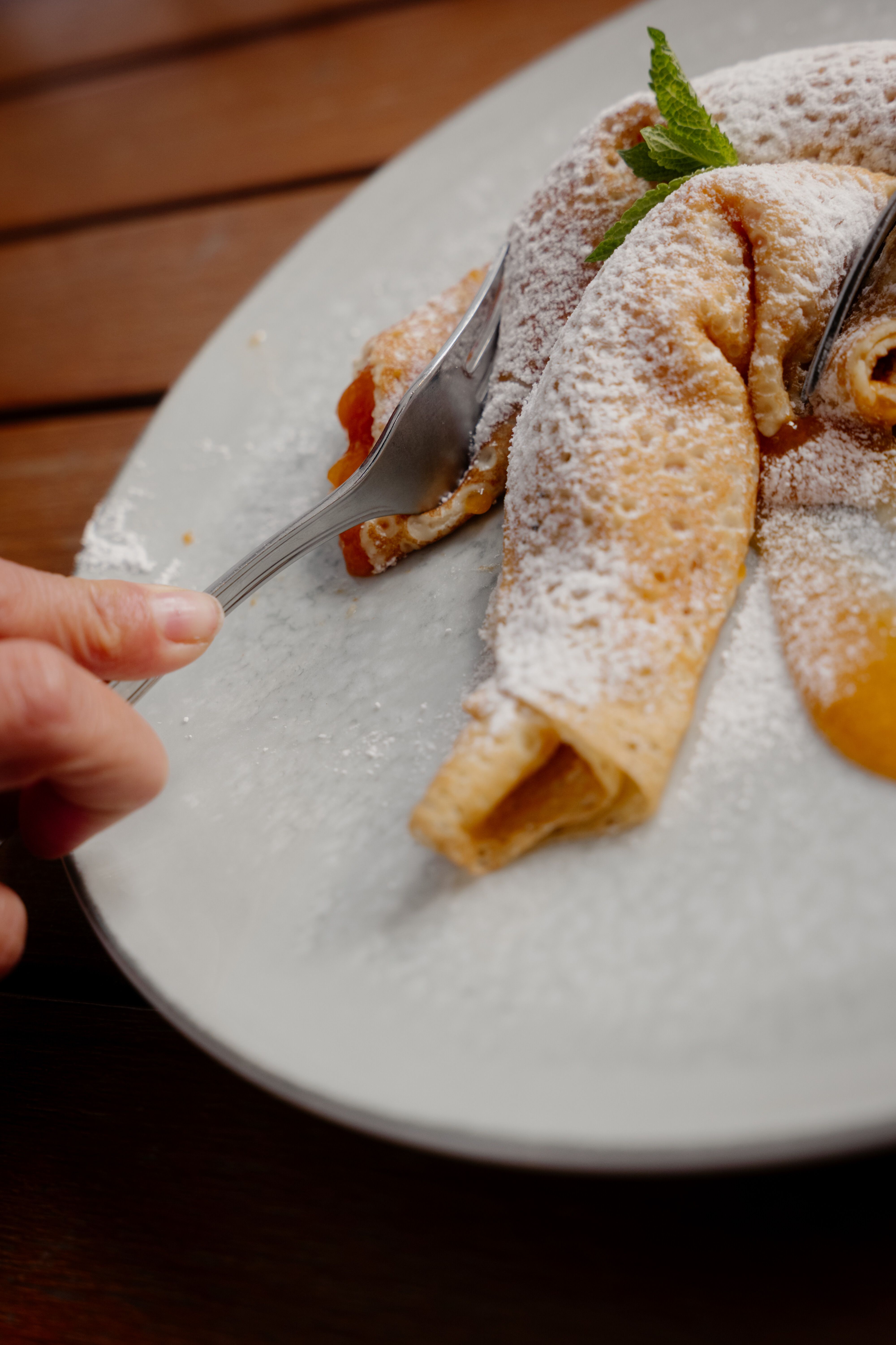 Gabel schneidet in eine mit Marillen gefüllte Palatschinke auf einem weißen Teller, bestäubt mit Staubzucker und mit Minzblatt garniert.