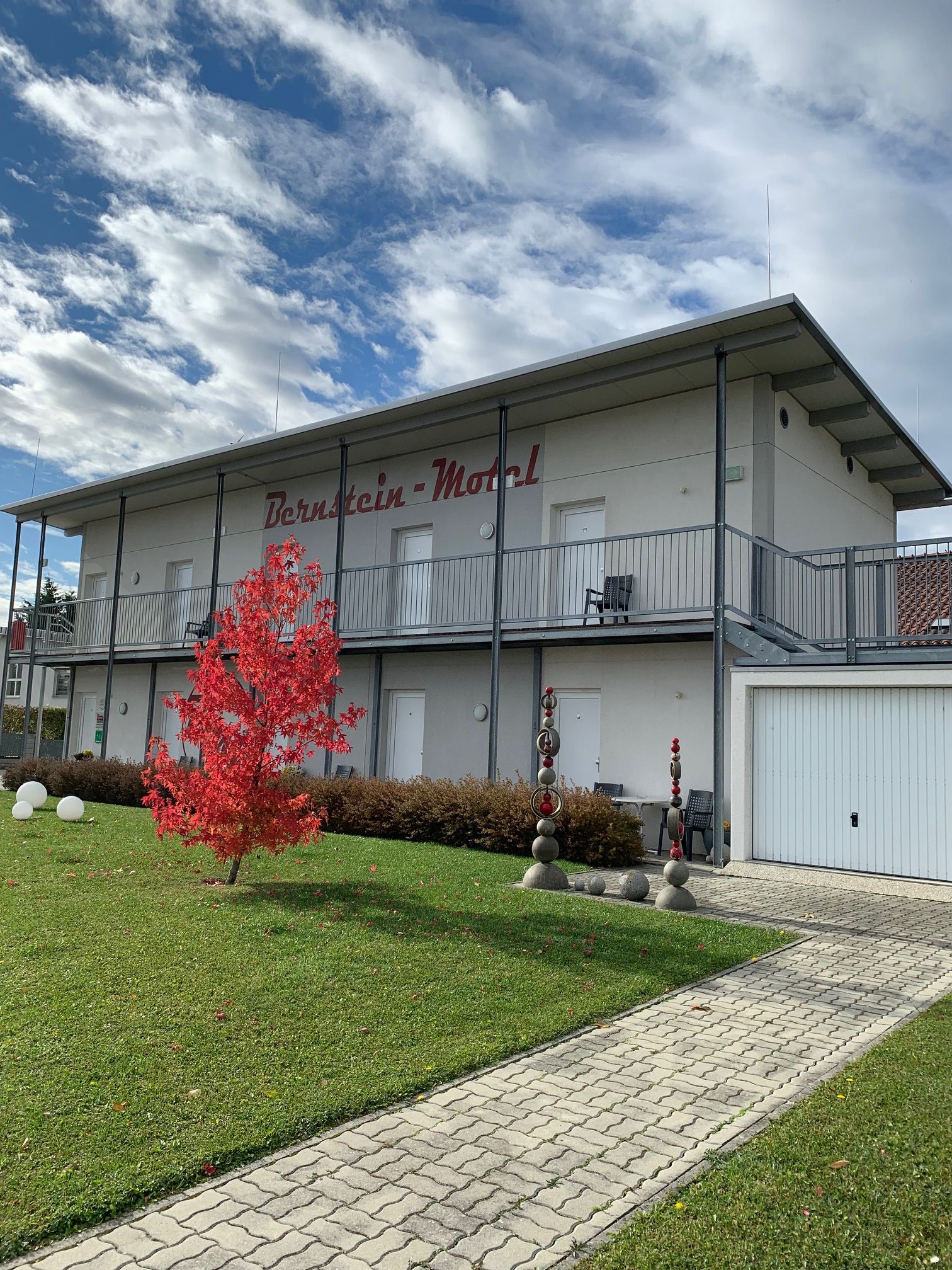 Zweistöckiges Gebäude mit der Aufschrift 'Bernstein Motel', roter Baum im Vordergrund, blauer Himmel mit Wolken.