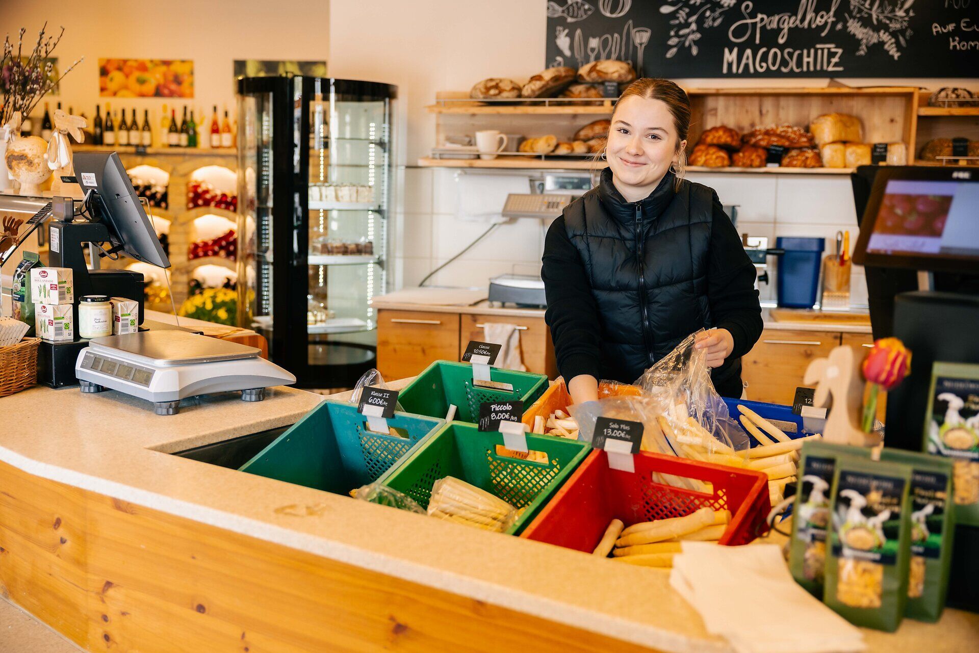 Innenaufnahme des Ab-Hof-Ladens Magoschitz. Eine Verkäuferin hinter dem Verkaufspult lächelt in die Kamera und packt Spargel ein.