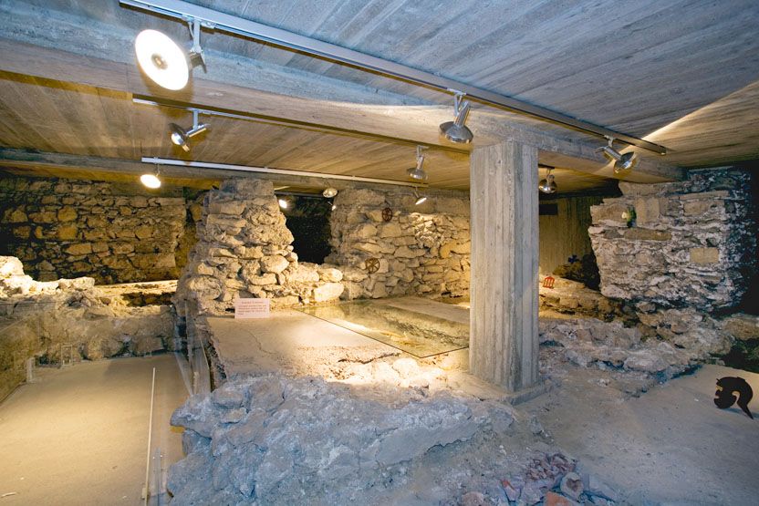 Unterirdische Ruinen mit Steinmauern und Betonstützen, beleuchtet durch Deckenstrahler.