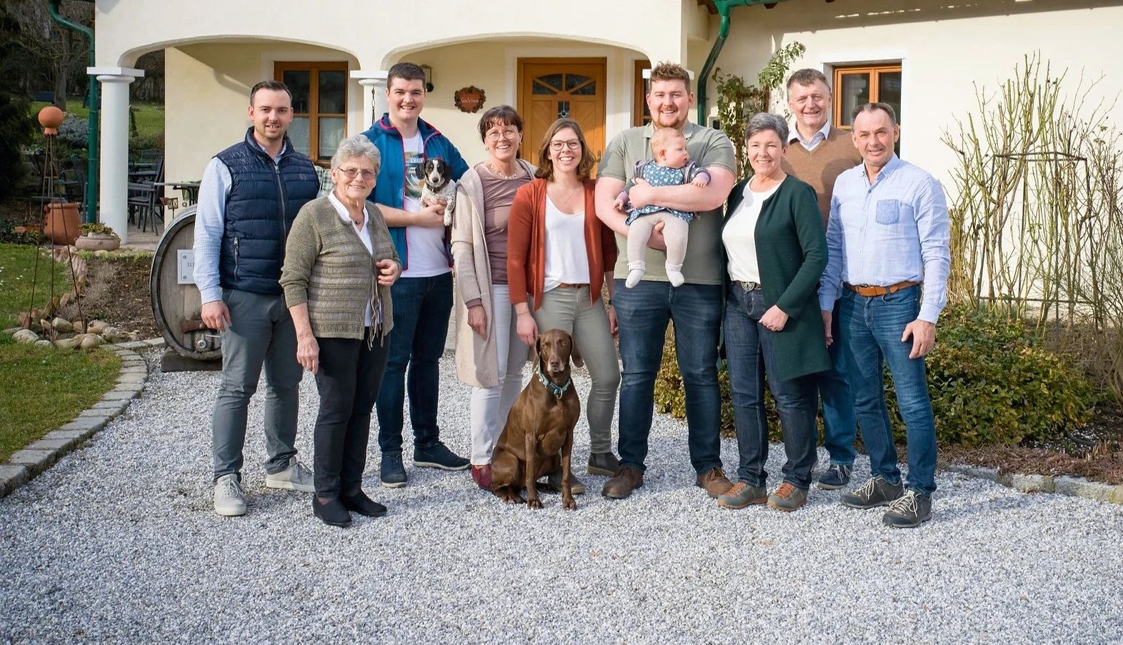 Eine Familie steht vor einem Haus, vier Generationen zusammen mit einem Hund und einem Baby.