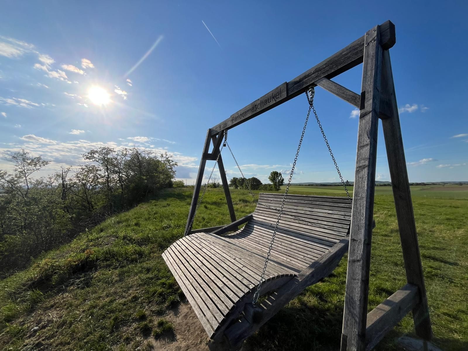 Große hölzerne Panoramaschaukel auf einer grünen Wiese unter strahlend blauem Himmel. Die tief stehende Sonne beleuchtet die Landschaft und wirft lange Schatten, während einzelne Wolken und ein Kondensstreifen den Himmel durchziehen. Die Szene wirkt ruhig und einladend mit weitem Blick über Felder und Natur.