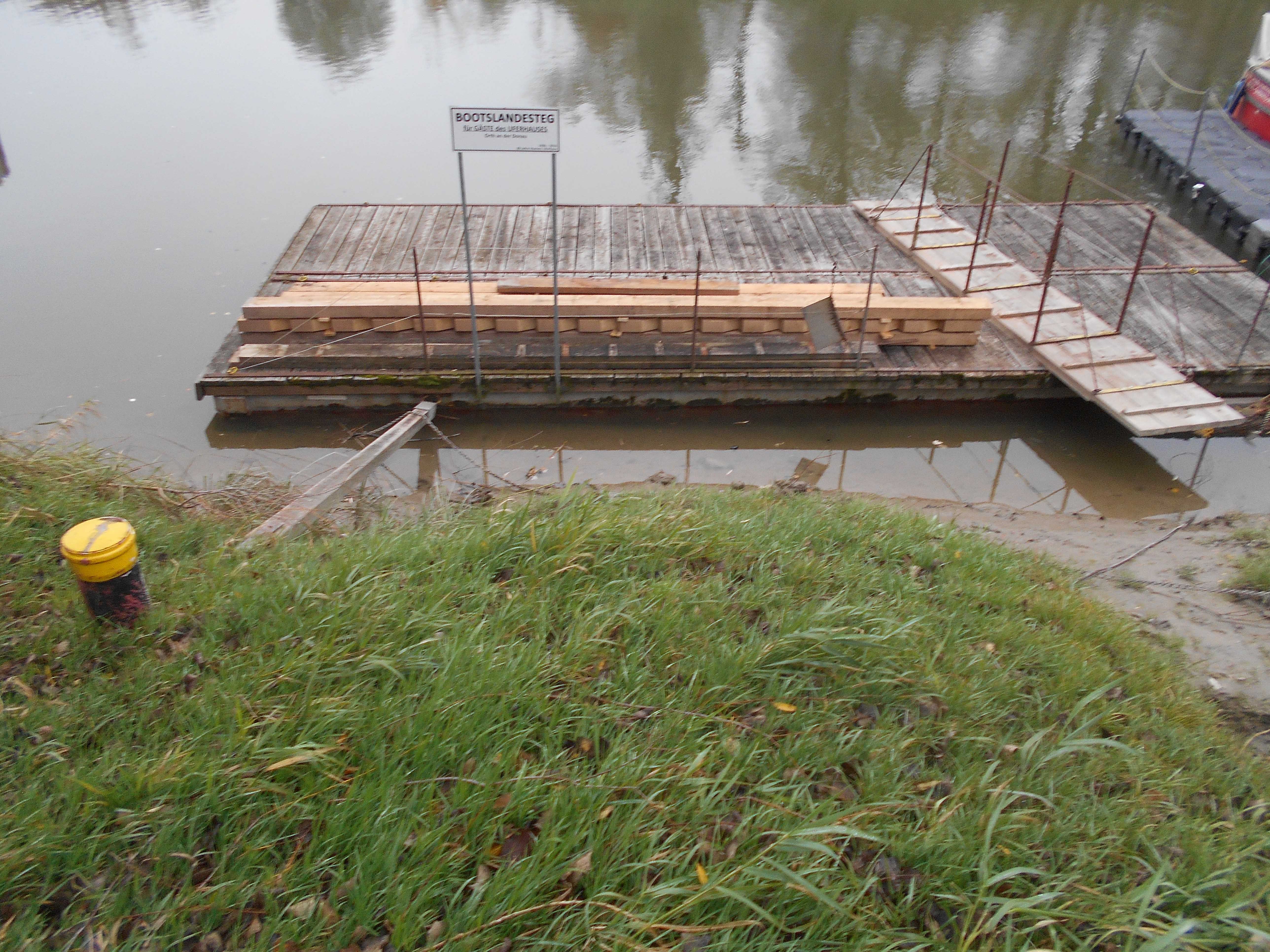 A wooden jetty with a sign and stacked wood, surrounded by water and grass.