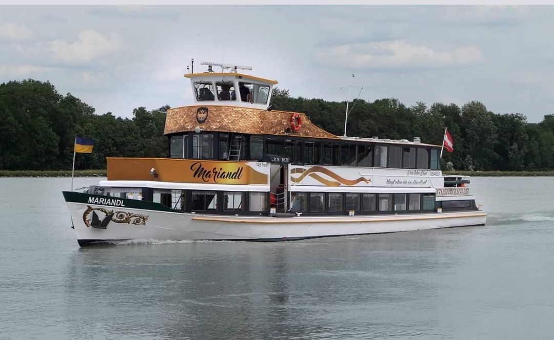 Ein Ausflugsschiff namens MS Mariandl fährt auf einem Fluss, umgeben von Bäumen. Es trägt die Flaggen der Ukraine und Österreichs.