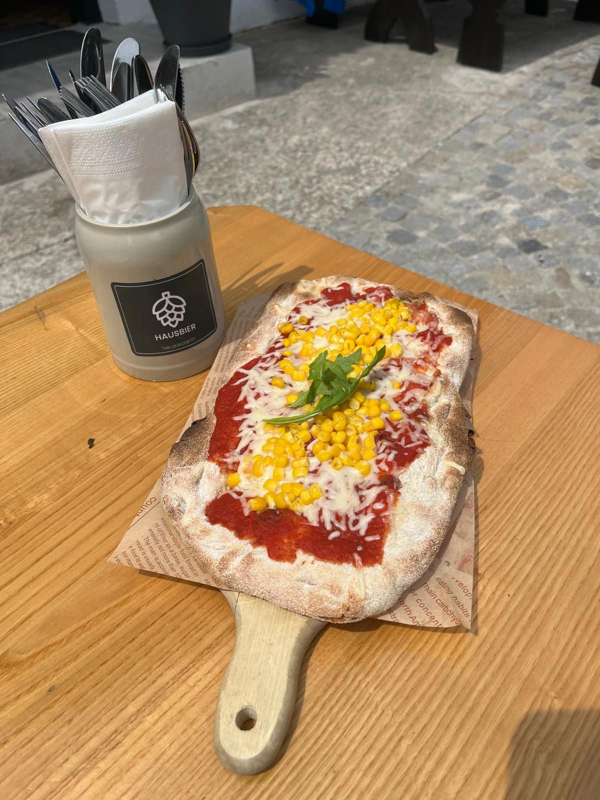 Fladen mit Mais und Rucola auf einem Holzbrett serviert, daneben ein Behälter mit Besteck und Servietten.