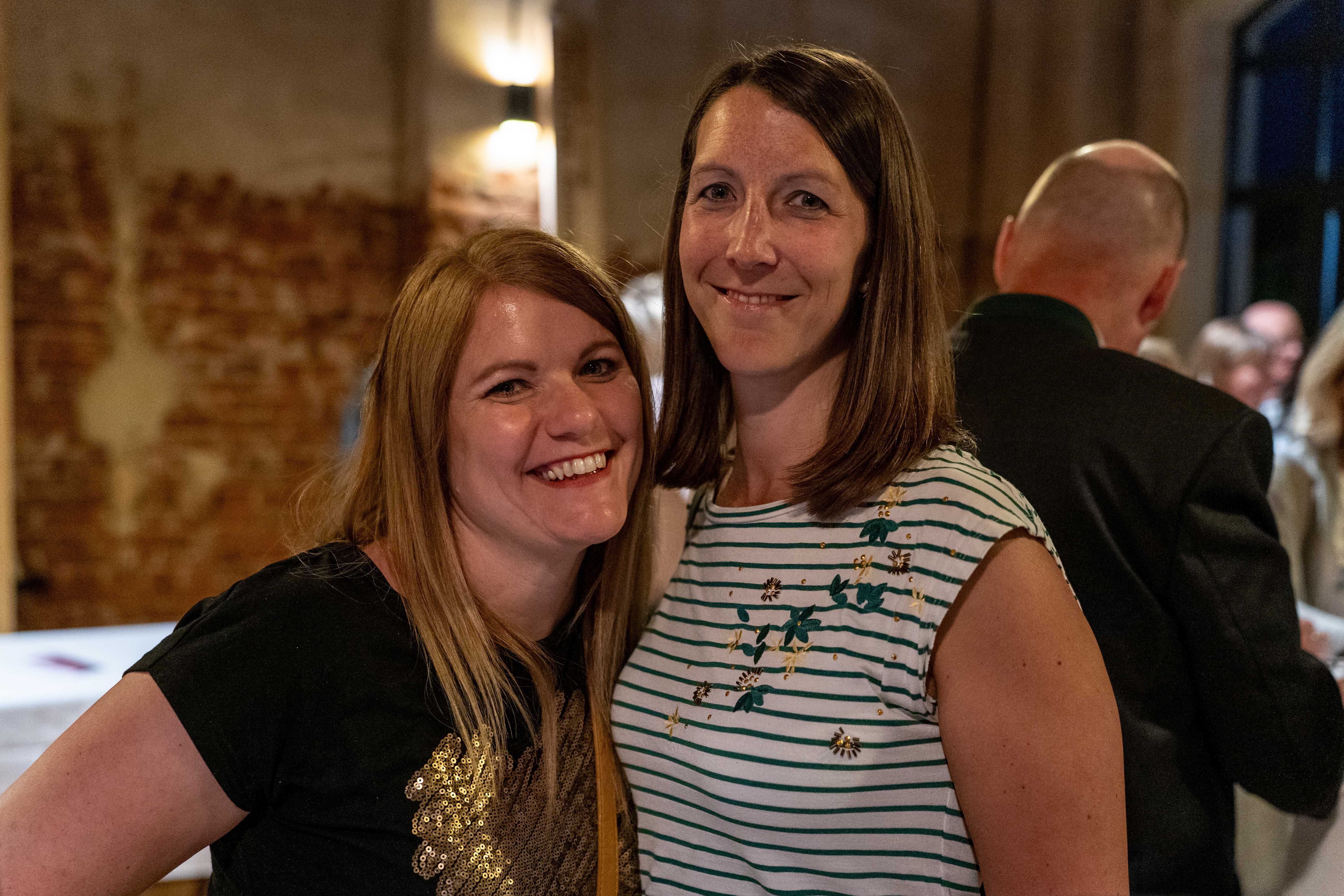 Zwei lächelnde Frauen posieren bei einer Weinveranstaltung in einer rustikalen Eventlocation mit Backsteinwand und Gästen im Hintergrund.
