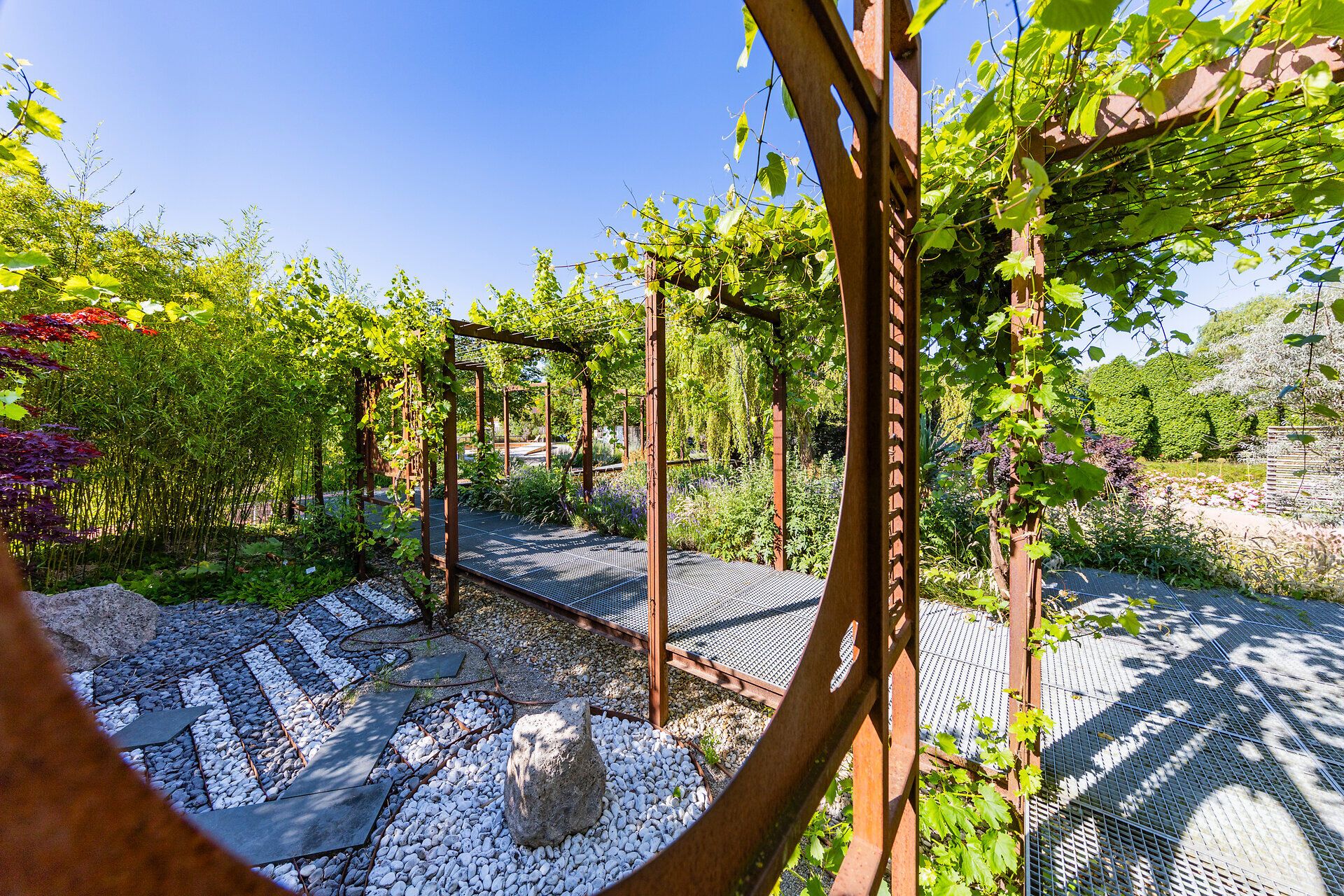 Ein malerischer Gartenweg schlängelt sich durch üppiges Grün, umgeben von blühenden Pflanzen und schattenspendenden Pergolen. Die harmonische Verbindung von Natur und Gestaltung lädt zum Verweilen und Entspannen ein, während die sanfte Brise die Blätter zum Flüstern bringt.