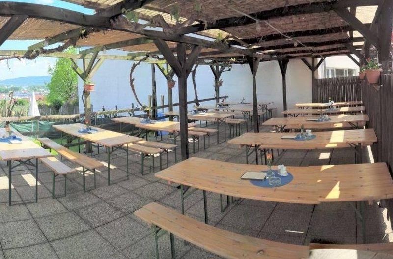 Leere Heurigenterrasse mit Holzbänken und Tischen unter einer Pergola.