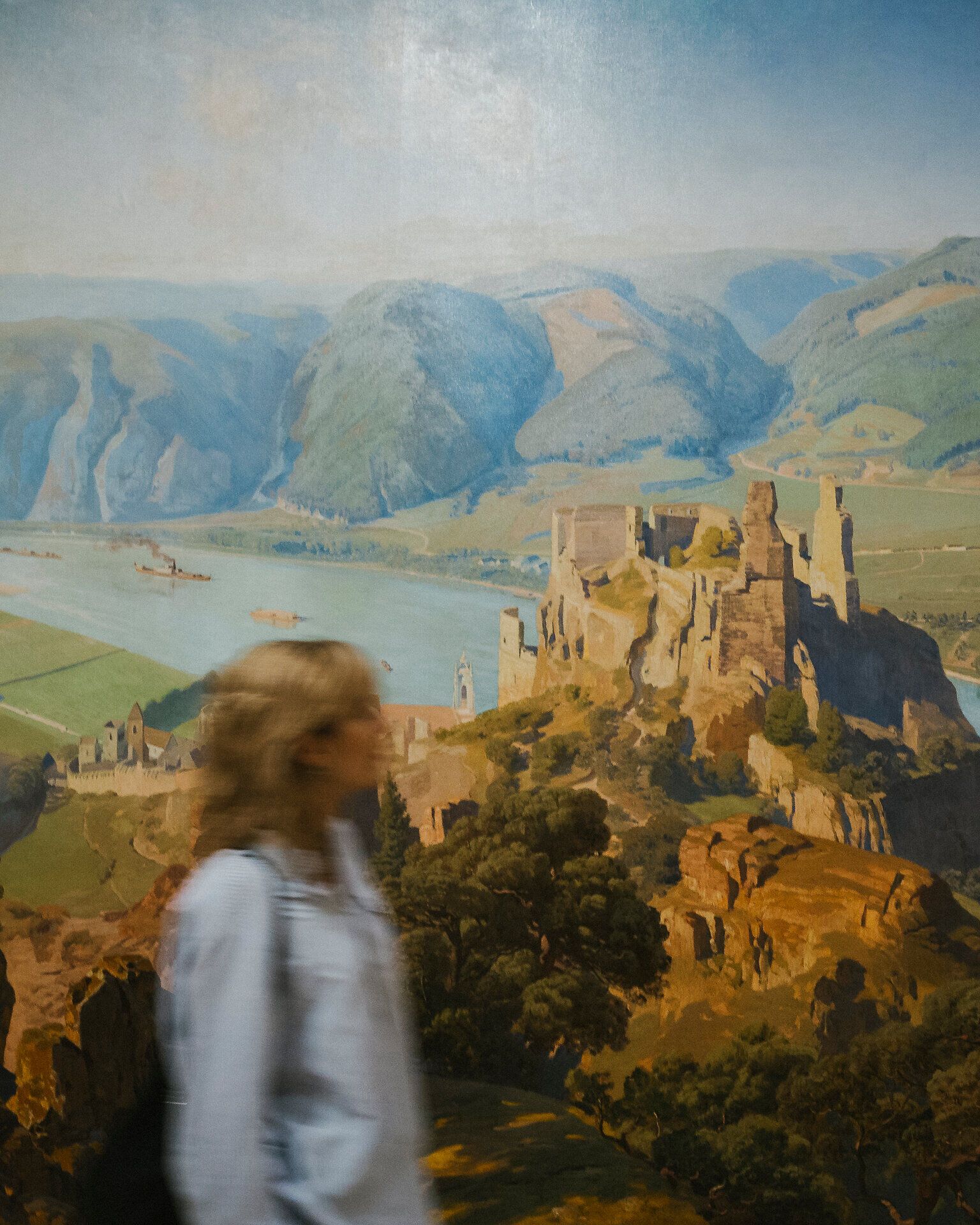 Unscharfe Person mit Rucksack geht vor einer großformatigen Wandmalerei mit Fluss, Hügeln und einer Burgruine auf einem Felsen vorbei.