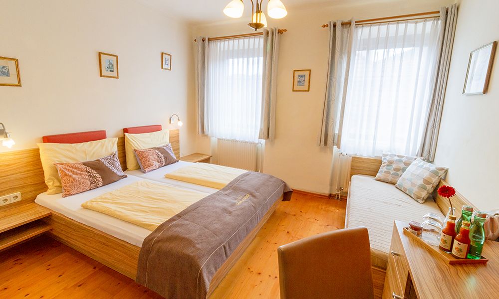 Ein gemütliches Gästezimmer mit Doppelbett, Tagesbett, Holzmöbeln und dekorativen Kissen. Helle Vorhänge und Bilder an den Wänden.