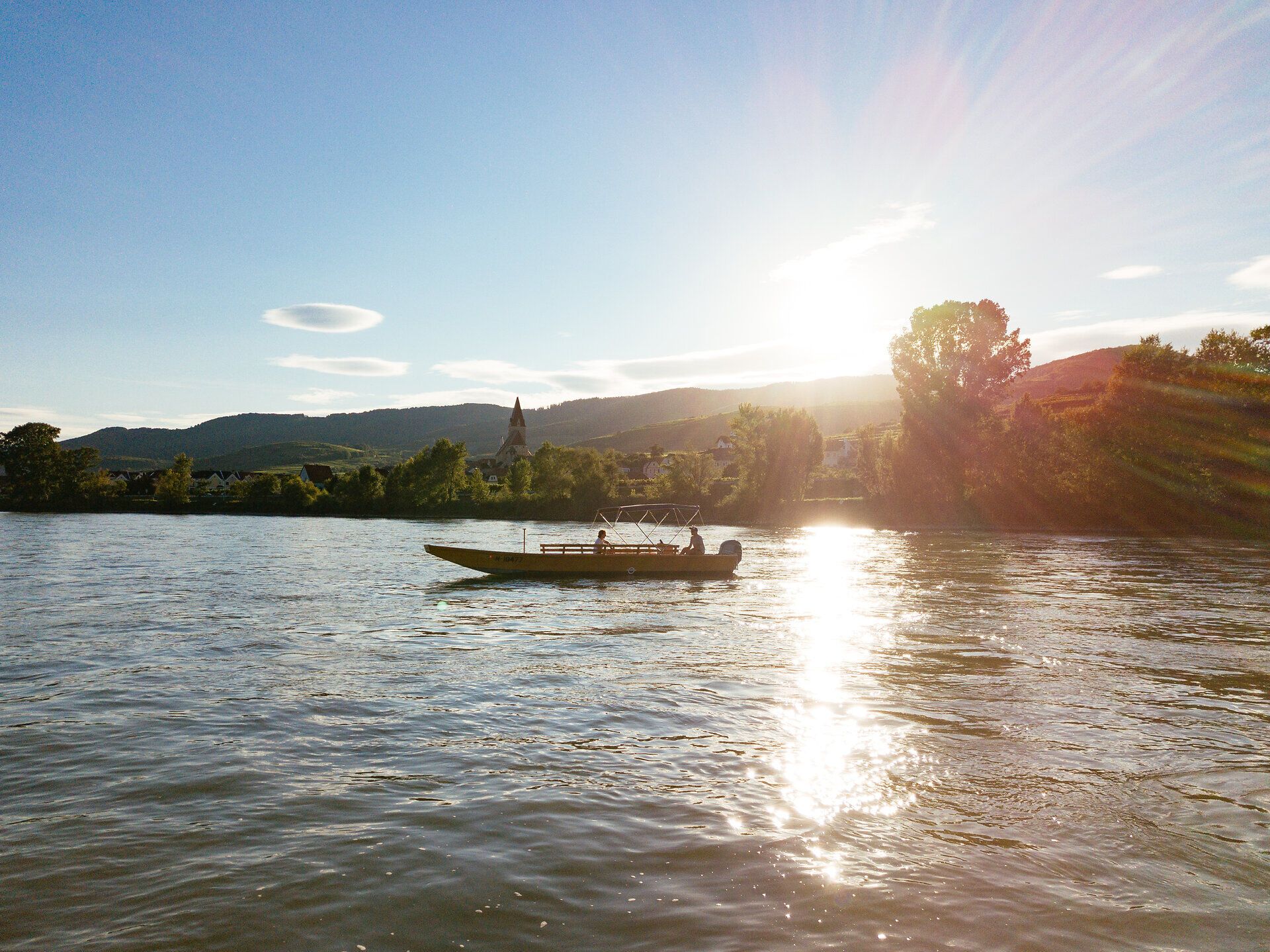Die sanften Wellen der Donau spiegeln das goldene Licht der untergehenden Sonne wider, während ein Boot gemächlich über das Wasser gleitet. Umgeben von üppigen grünen Ufern und sanften Hügeln, bietet diese Bootstour eine perfekte Gelegenheit, die atemberaubende Landschaft der Wachau zu genießen.