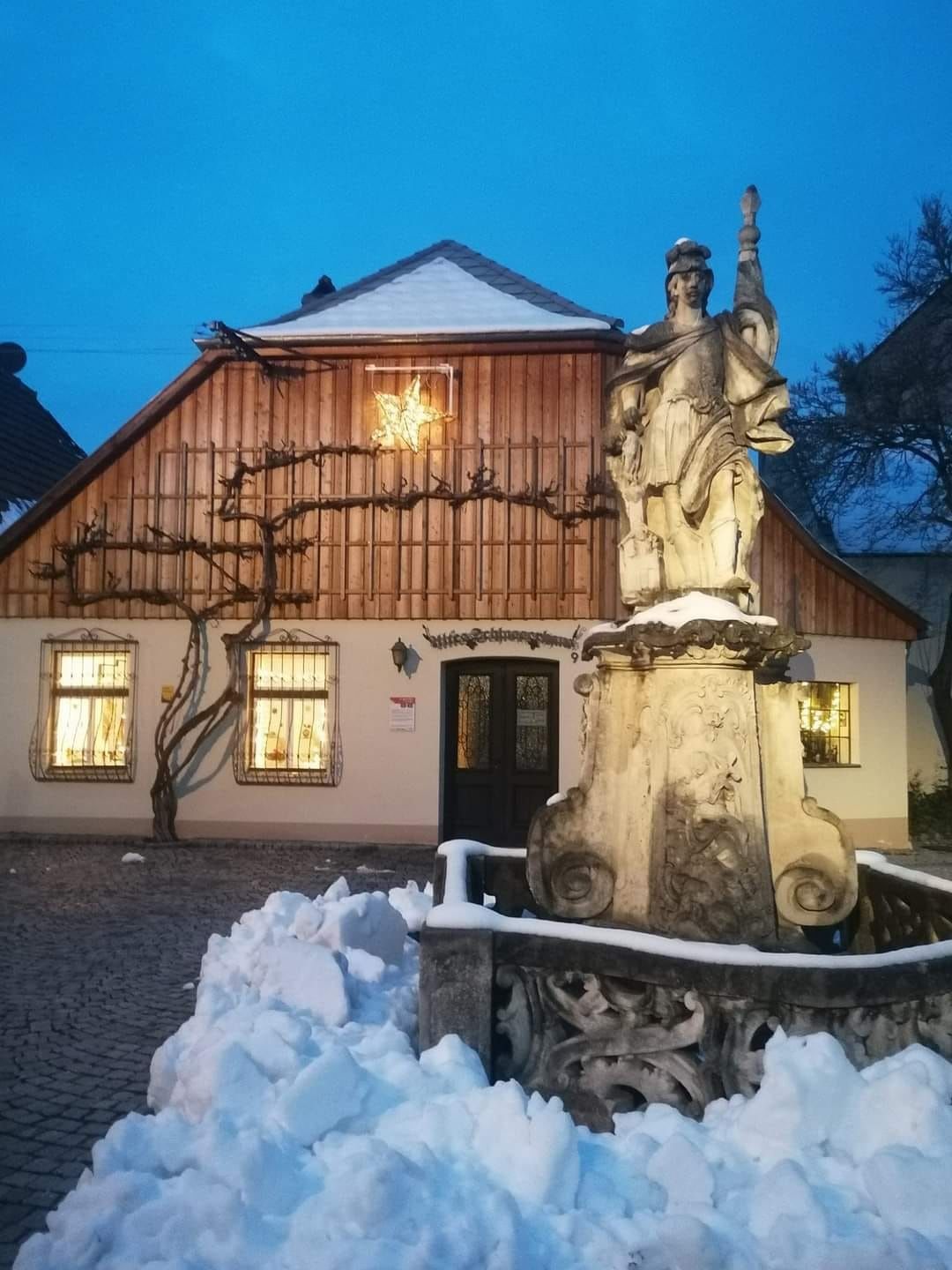 Ein verschneites Haus mit einer Statue im Vordergrund, beleuchtet von einem Stern an der Fassade.