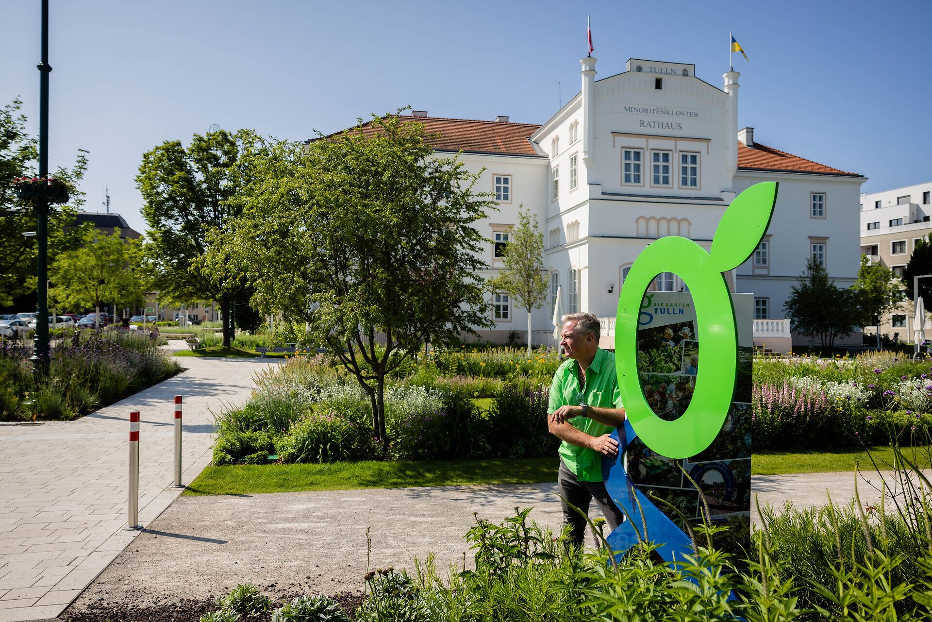 Ein Gärtner in Tulln steht vor dem Rathaus neben einem Symbol der GARTEN TULLN. Es sind blühende Sträucher und Bäume zu sehen, blauer Himmel und Sonnenschein.