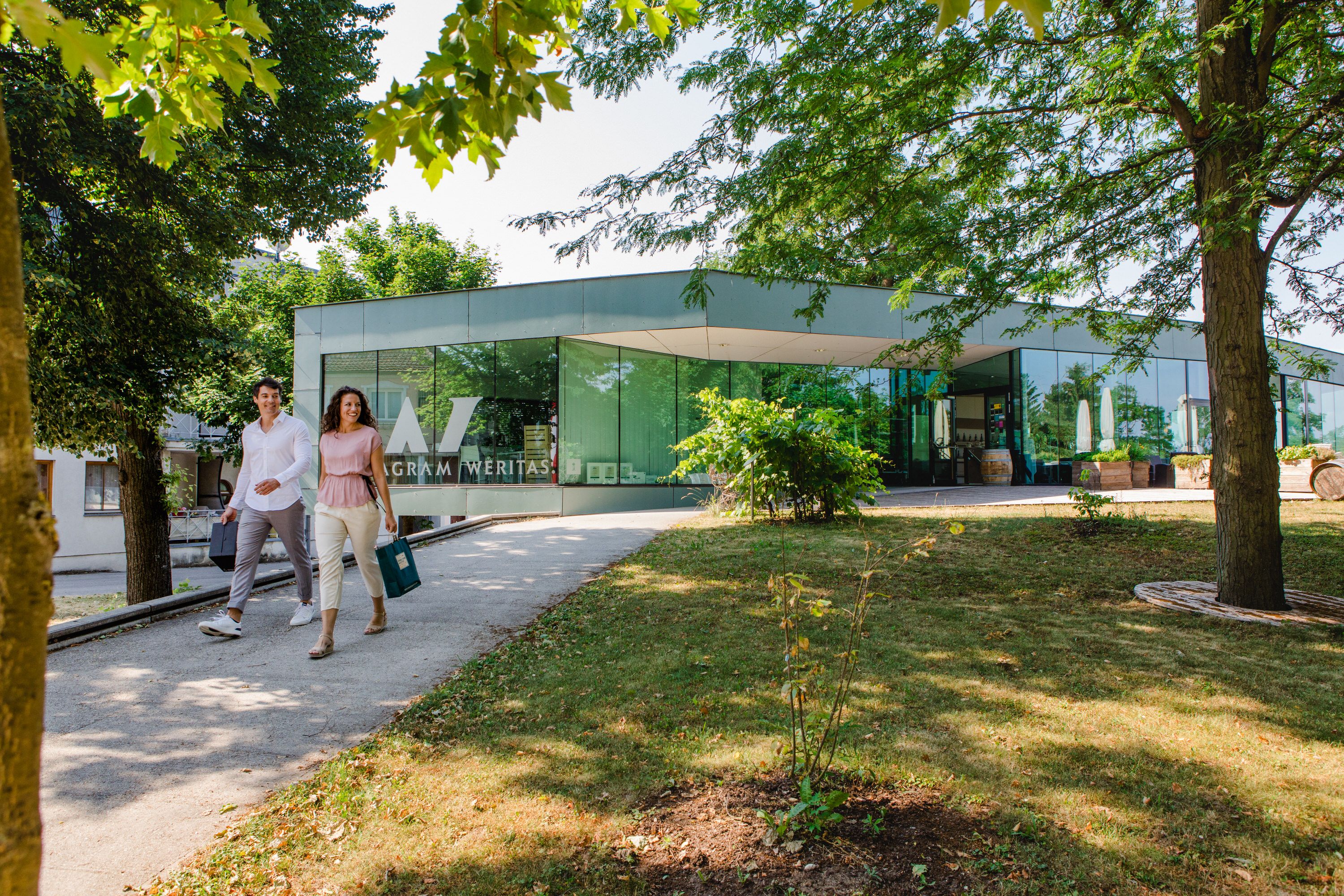 Personen spazieren auf einem schattigen Weg vor der moderen Gebietsvinothek  Grünen.