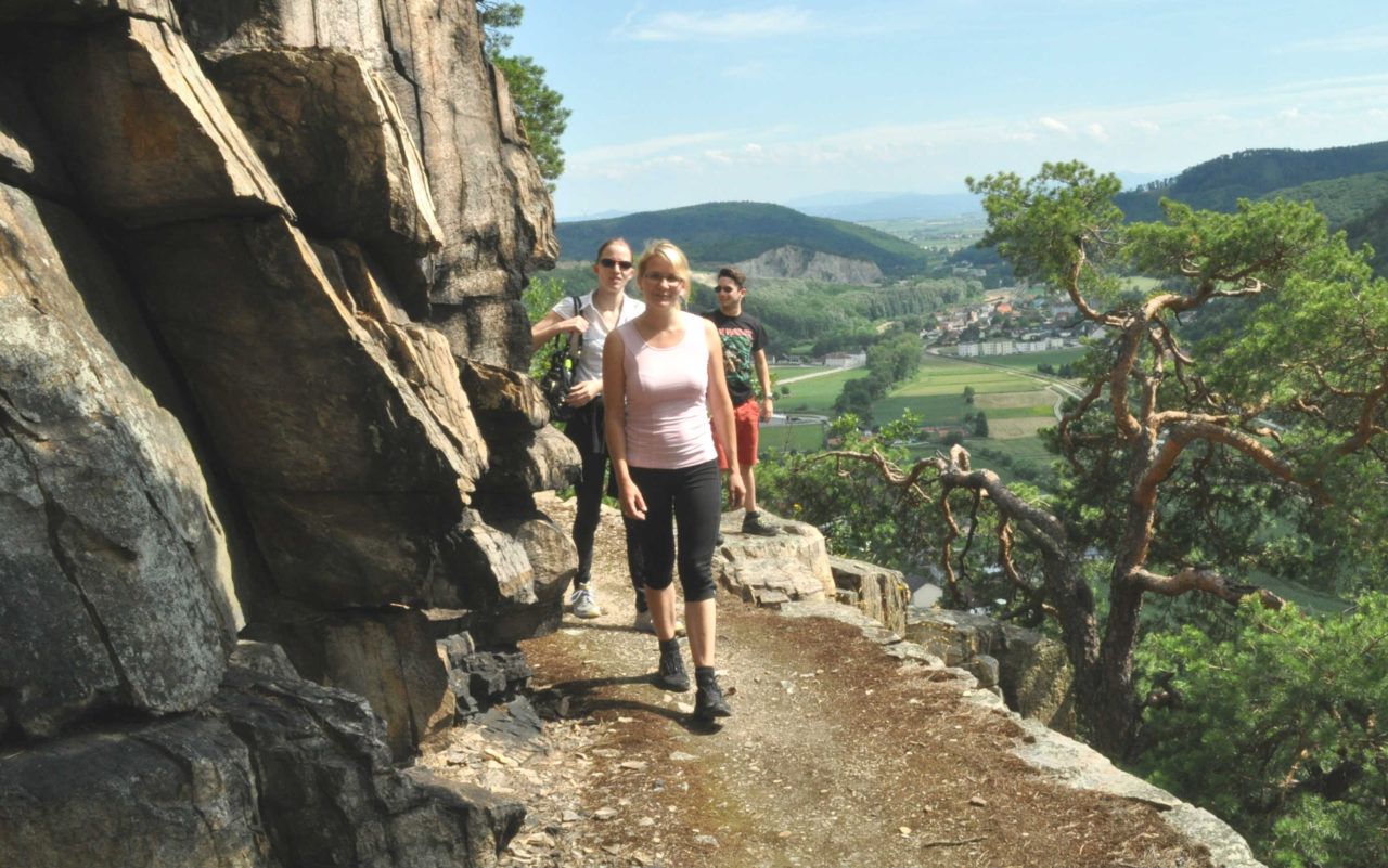 Drei Personen wandern auf einem schmalen Pfad entlang einer Felswand mit Blick auf eine grüne Landschaft.