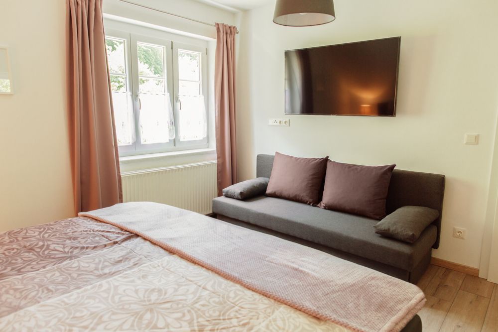 Innenansicht eines Schlafzimmers mit Bett, Sofa, Fenster und Wandfernseher.