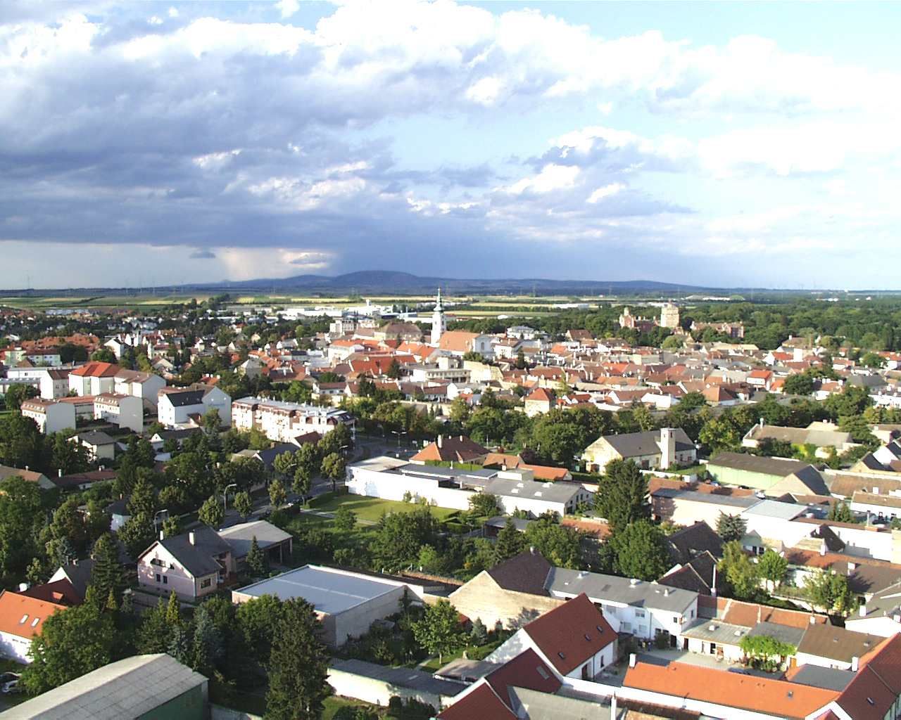 Luftaufnahme von Bruck an der Leitha mit Häusern und Kirche, umgeben von grüner Landschaft und bewölktem Himmel.