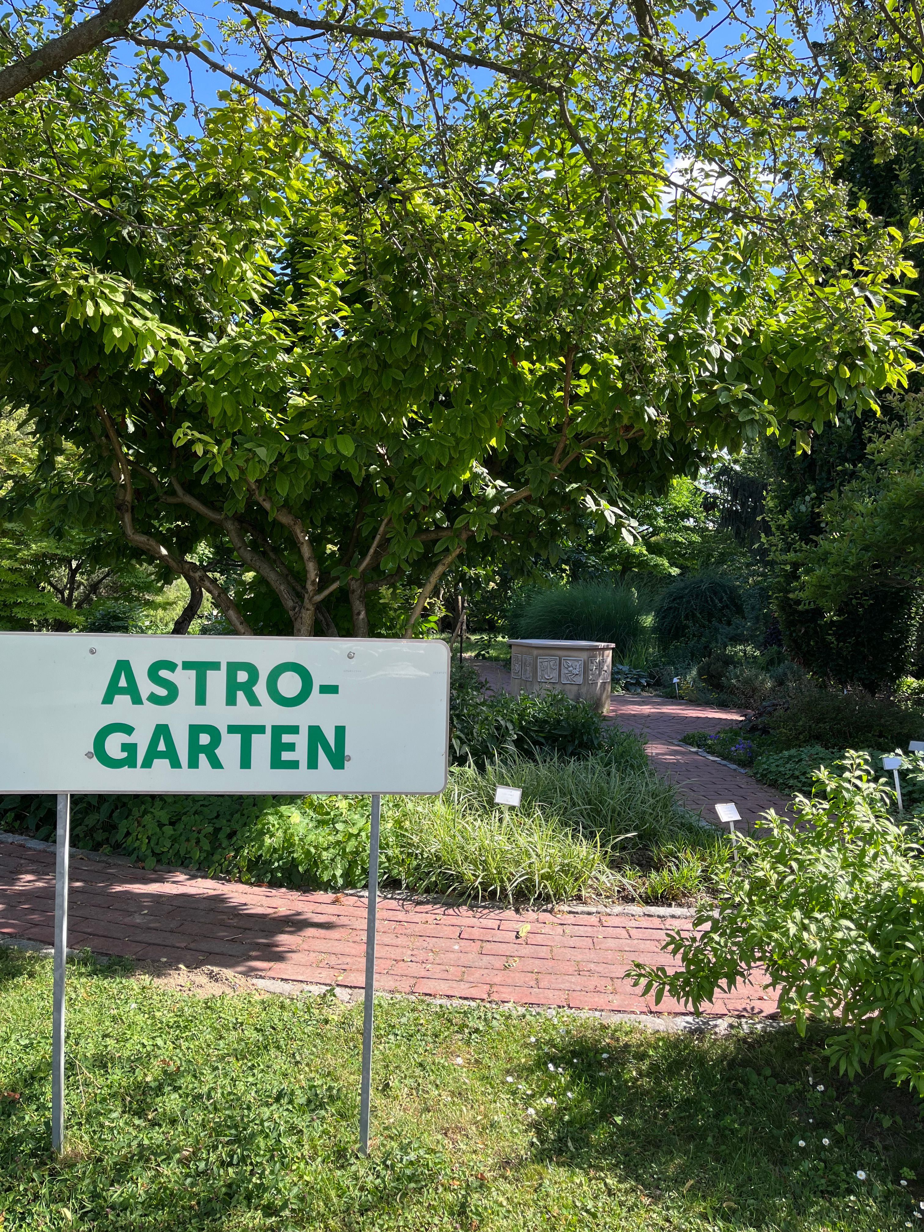 Ein Schild mit der Aufschrift 'Astro-Garten' steht vor einem grünen Garten mit Bäumen und einem gepflasterten Weg.