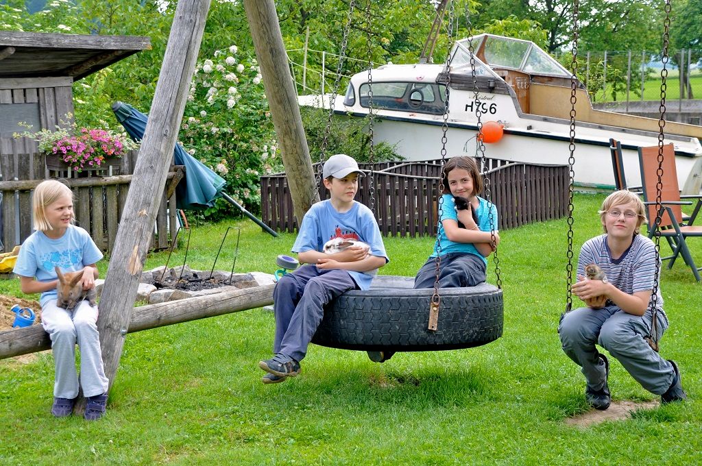 Vier Kinder sitzen im Garten auf einer Schaukel und halten Tiere. Im Hintergrund steht ein Boot.