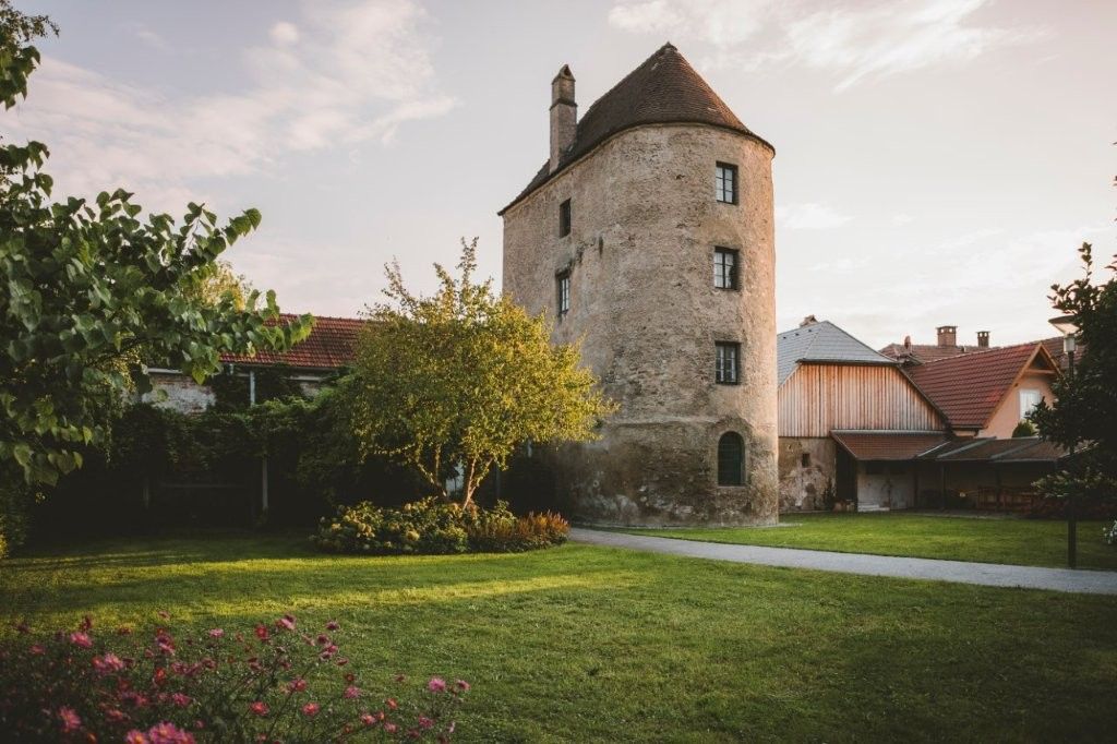 A medieval round tower in a well-kept garden filled with blooming flowers and trees, surrounded by buildings with red roofs.