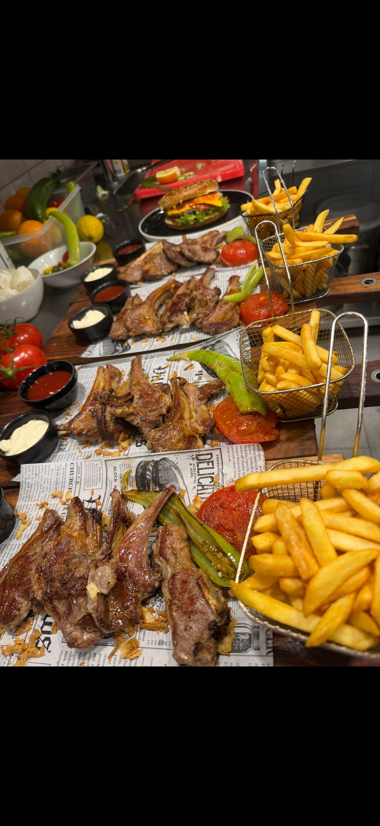 Egy asztal tele grillezett bárányszeletekkel, kosárban sült chipsekkel és friss zöldségekkel körülvett hamburgerrel.