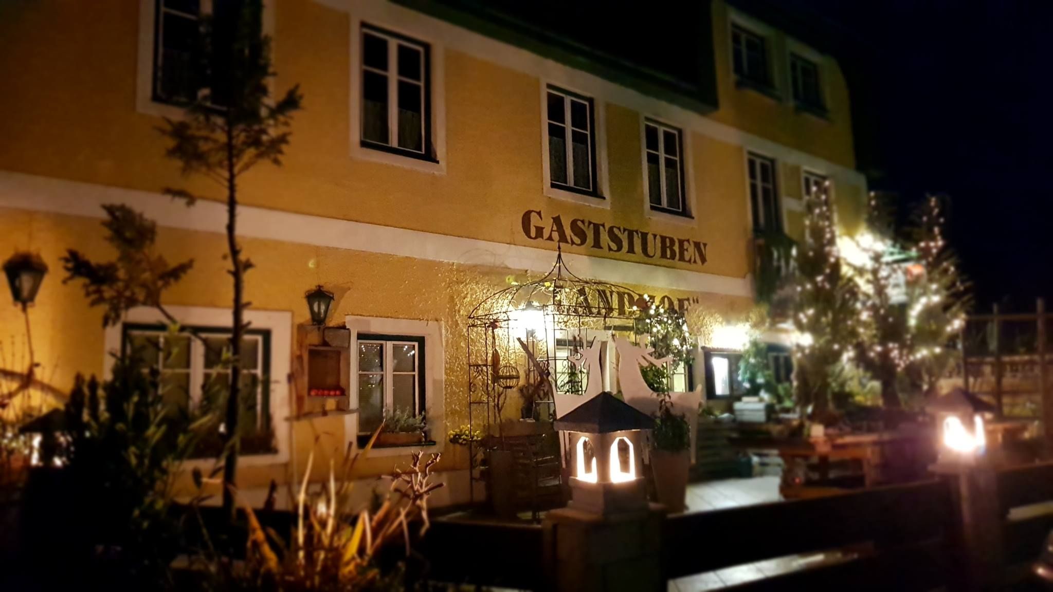 Nachtansicht eines gelben Gebäudes mit der Aufschrift 'Gaststuben Landhof', beleuchtet von Laternen und umgeben von Pflanzen.