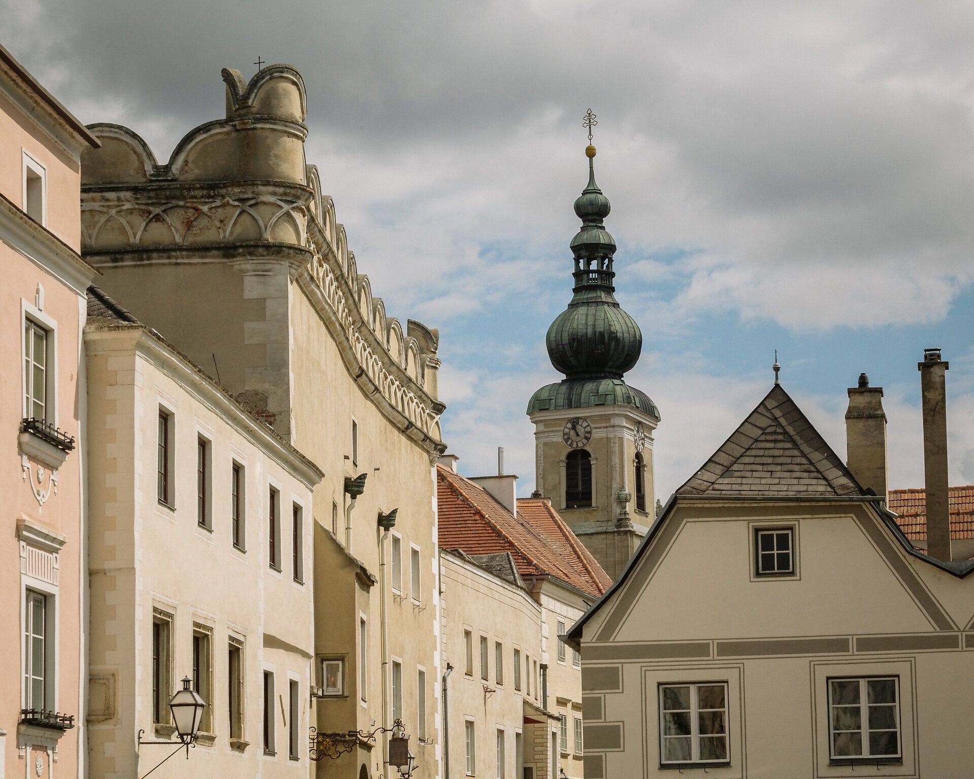 Blick entlang einer engen Straße mit historischen Fassaden und roten Dächern; im Hintergrund ragt ein barocker Kirchturm mit Zwiebelhaube vor grauem Himmel auf.