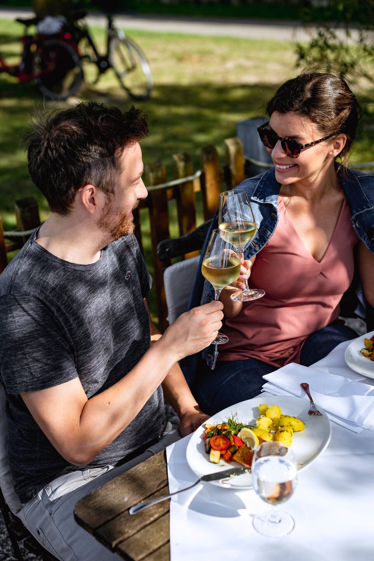 Ein Paar sitzt im Gastgarten und stößt beim Essen mit Gläsern Weißwein an.